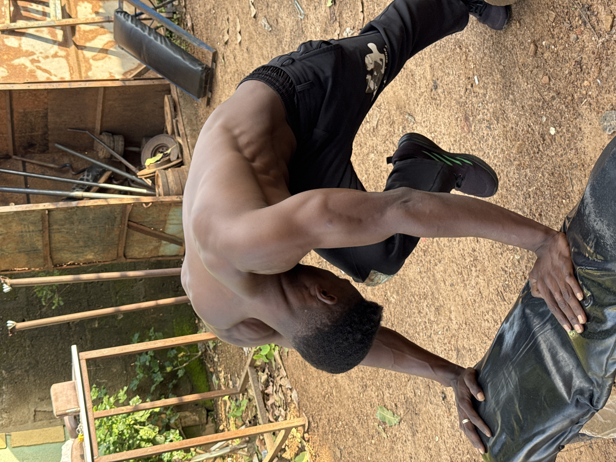 The image depicts a shirtless Black male in athletic attire, performing an exercise on a black padded surface, which appears to be part of a weight bench or similar gym equipment. He is wearing black track pants with green stripes on his sneakers. The man's physique is muscular, with visible abdominal and back muscles. His hands are placed on the padded surface, and his body is arched backwards.
The background suggests an outdoor or semi-outdoor setting, possibly a makeshift gym or an informal training area. There are metal structures, including a rack-like frame and what appears to be part of a corrugated metal shed or wall. Weights and other gym equipment are visible in the background, scattered around the area. The ground is a mix of dirt and gravel, with some dry leaves and debris. The lighting indicates it is daytime, with sunlight casting shadows. The overall environment appears somewhat rustic and worn.
The image is believed to have been taken in the city of Akwanga, Nigeria.