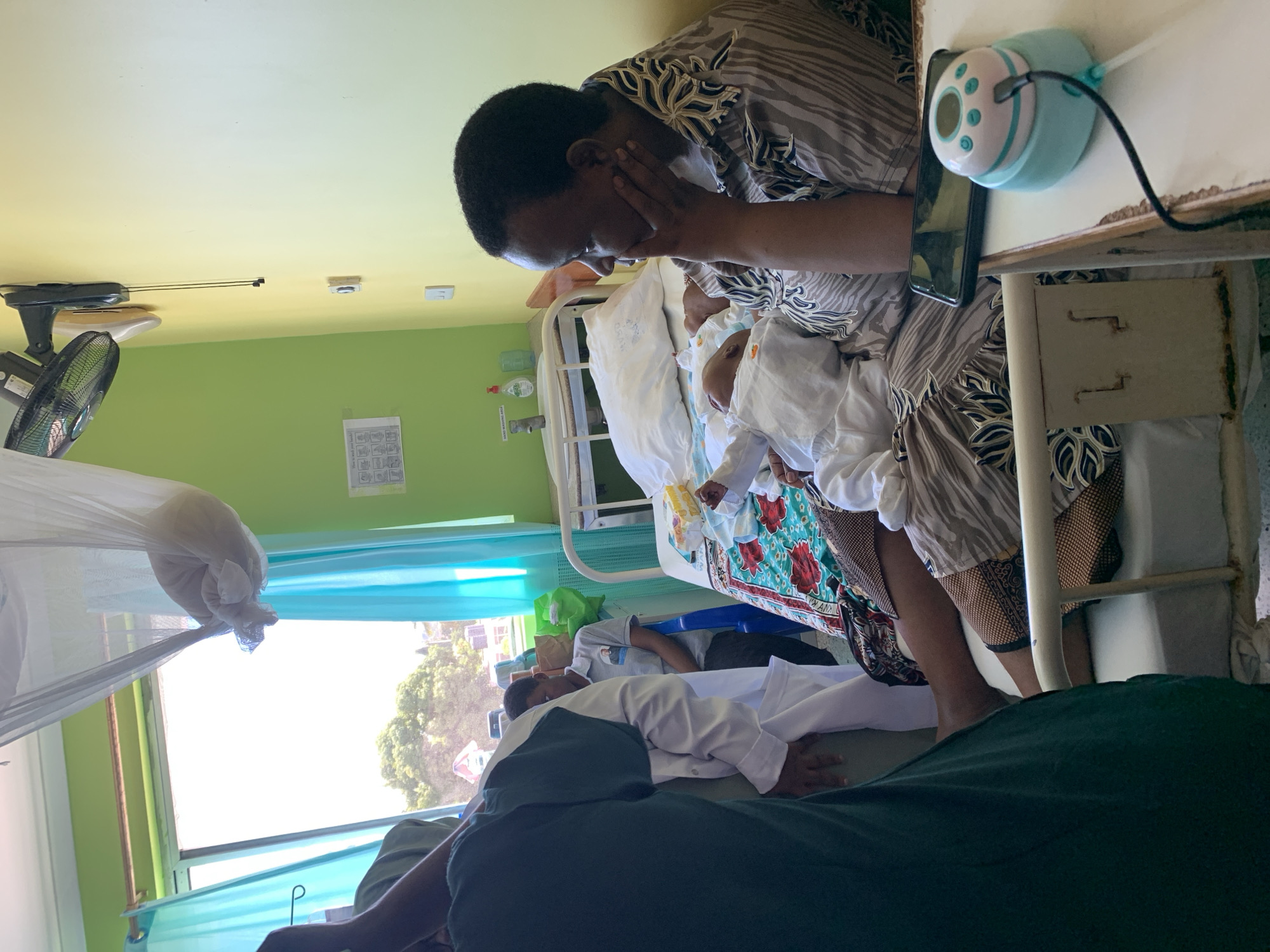 A woman in a patterned shirt sits by a hospital bed holding a baby in white clothing. Her hand is to her face, and she appears to be looking down at the infant. On the bed table next to her, there is a smartphone and a light blue device with buttons. Another person lies prone on a nearby bed. The room has green walls, a fan, and a mosquito net. Based on the image's background and context, it is presumed that this photo was taken in a hospital in the city of Dar es-Salaam, Tanzania.