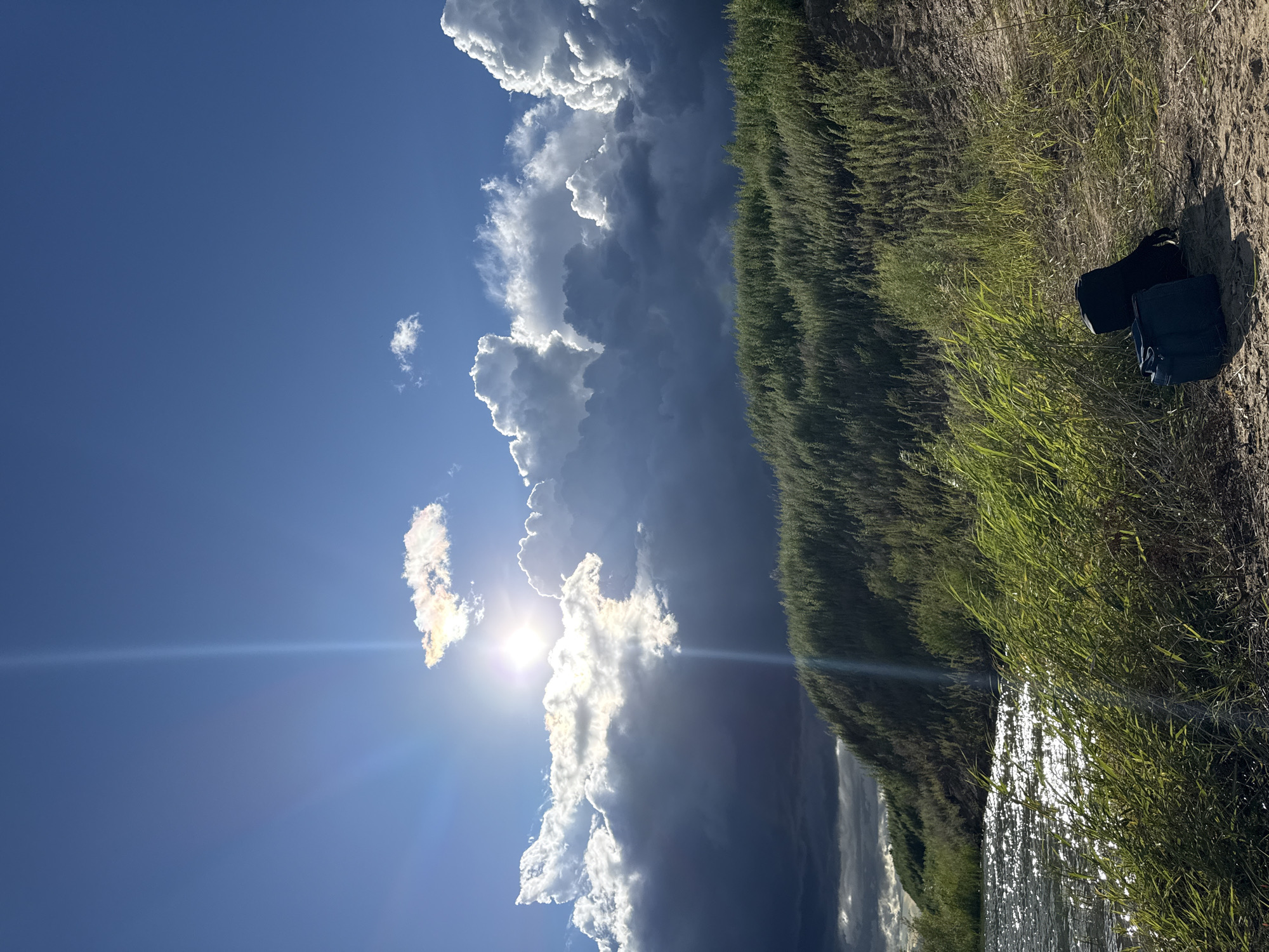 The image depicts a natural landscape with a body of water in the foreground, bordered by a grassy embankment. The sky is partly cloudy with the sun prominently visible, casting rays of light. Two bags, one black and one blue, are placed on the sandy ground near the grass. The environment suggests a rural or undeveloped area, possibly a riverbank or lakeside. The specific location in Blagoveshchensk, Russia, is not visually ascertainable from the image itself. There are no people or discernible actions occurring in the scene.