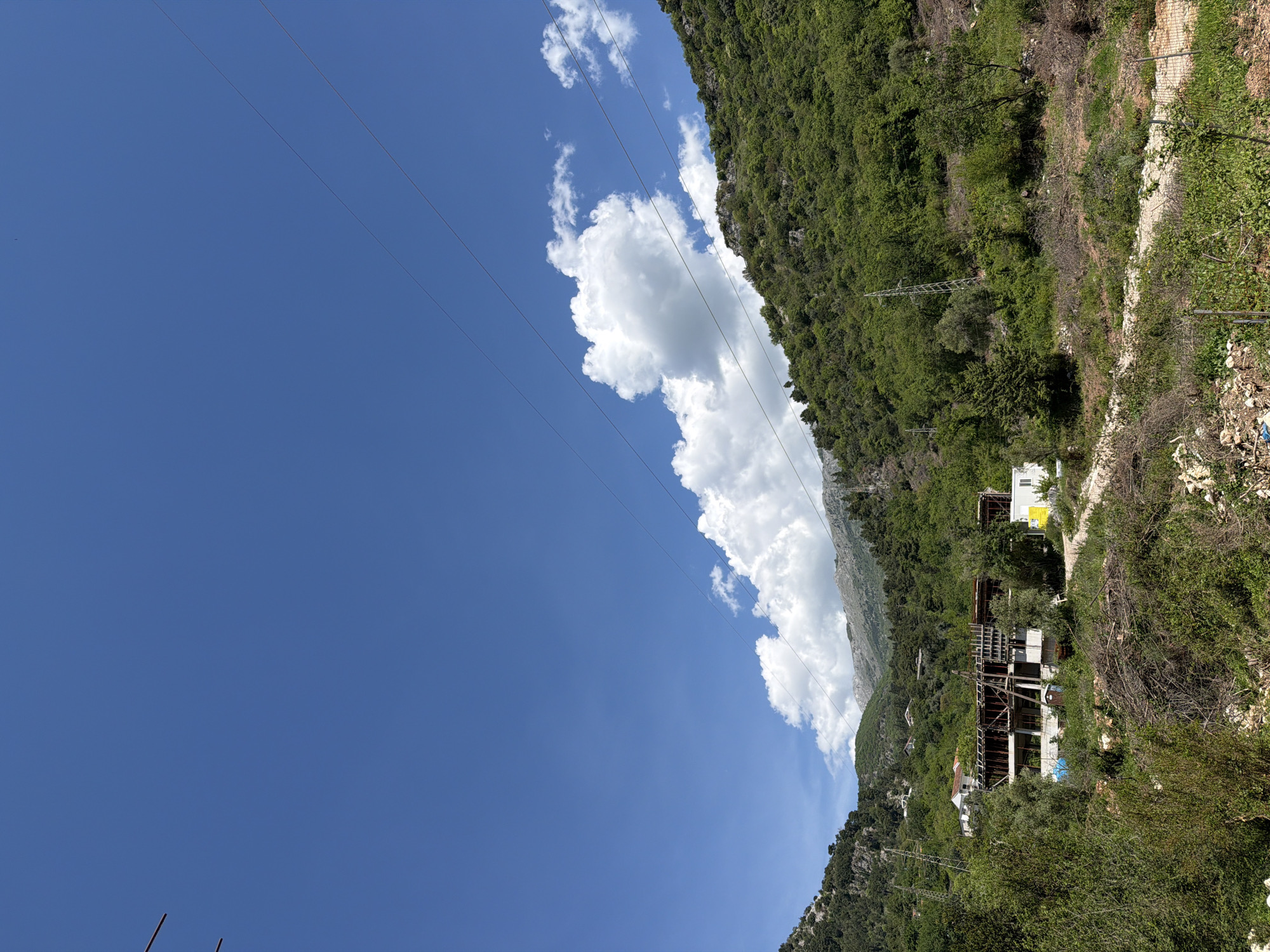 A vibrant outdoor scene in Budva, Montenegro, presents a mountainous landscape under a bright blue sky. The upper portion of the image is dominated by a clear, azure sky punctuated by several white, fluffy cumulus clouds, one of which forms a large, distinct mass. Multiple thin power lines traverse the sky from left to right, implying an overhead electrical grid.
The midground and background are characterized by steep, verdant hills and mountains, densely covered in rich green trees and vegetation. A prominent metal lattice power pylon stands on one of the hillsides, blending with the foliage.
Human presence is evident on the right side of the image. Higher on the slope, a white building with a distinctive yellow section, possibly a sign or architectural feature, is visible. Below it, a significant multi-story structure appears either unfinished or abandoned, showcasing exposed concrete framework and structural elements. Further down, the foreground reveals a rough path or track bordered by a wire fence, with scattered bushes, dry vegetation, and some visible debris or waste.
The scene is illuminated by bright daylight, suggesting sunny weather. The overall impression is a blend of natural beauty and human development, with signs of construction or a past settlement amidst the lush Montenegrin landscape. On the yellow section of the upper white building, faint, unreadable text is present. No specific activity or individuals are depicted.