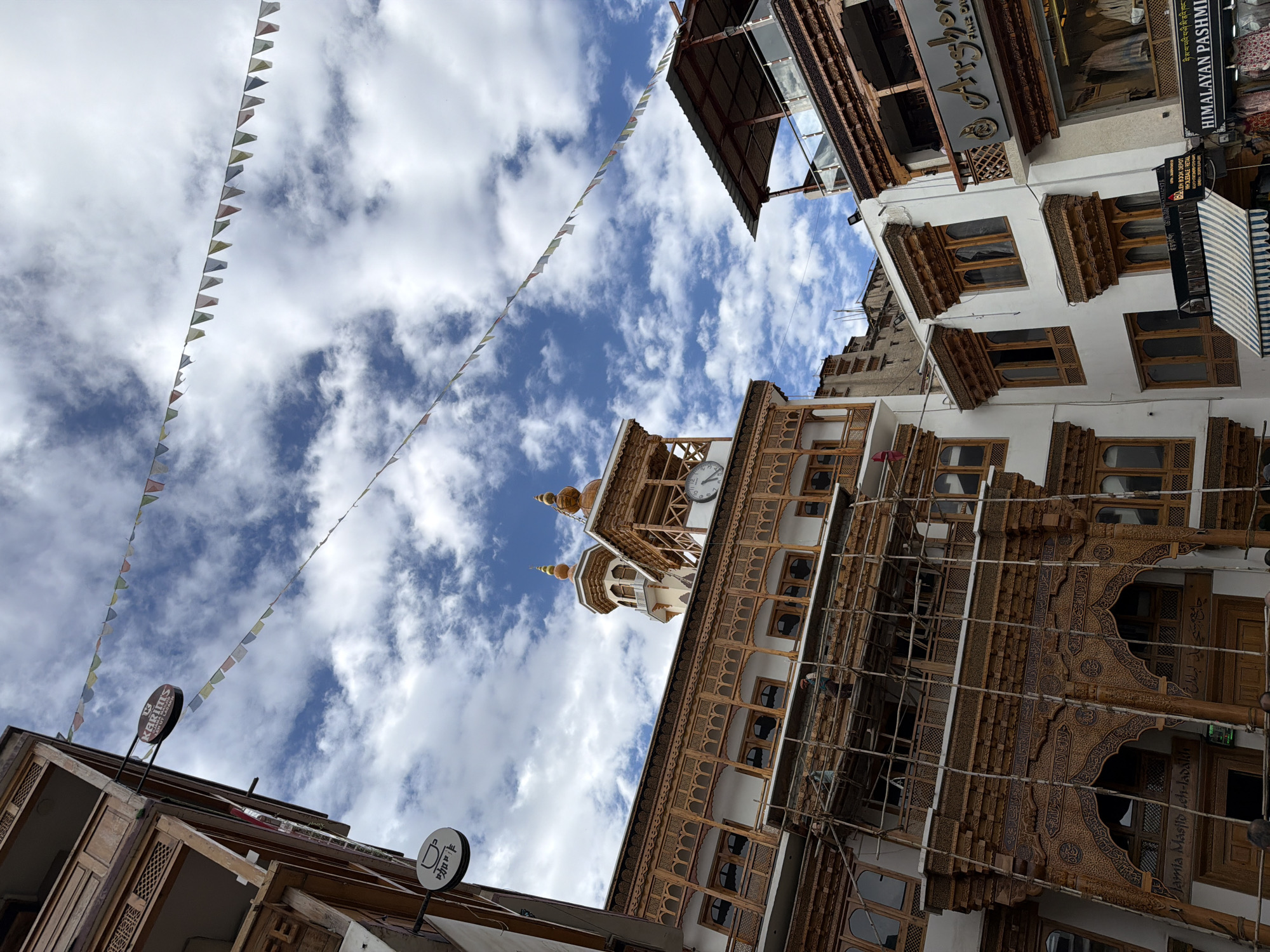 This image captures a street scene in Leh, India, featuring traditional Ladakhi architecture. The buildings showcase intricate wooden carvings and layered balconies. Prayer flags are strung across the sky, adding a vibrant touch to the cloudy blue expanse. In the foreground, a building displays ornate wooden arches with Islamic calligraphy, identified as Jamia Masjid. Scaffolding is present on one of the buildings, suggesting ongoing construction or maintenance. Signs for "Karim's" and "Arshon Fine D" are visible on shops. People are present on the ground level, some appearing to be shoppers. The overall atmosphere suggests a bustling urban environment with a strong cultural heritage.