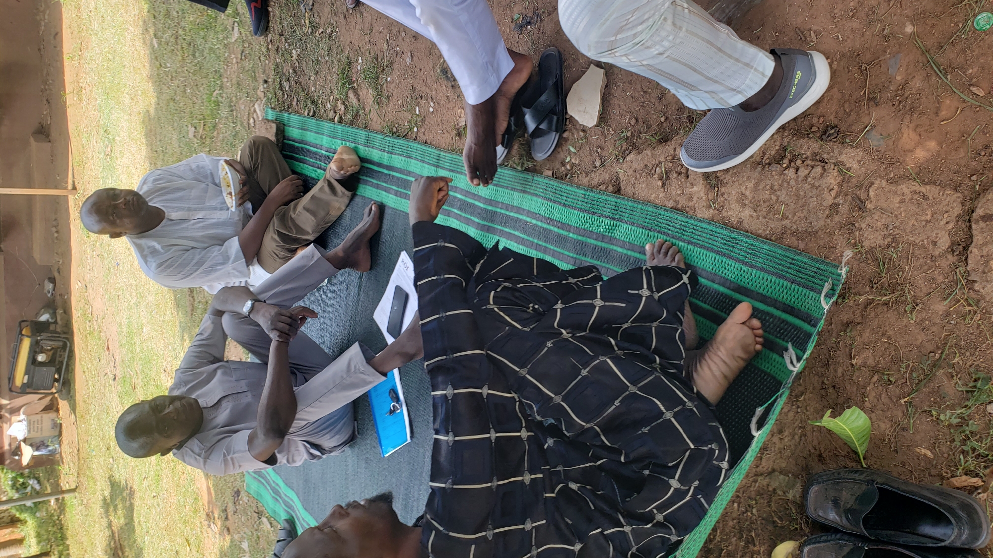 The image shows a group of men sitting on a green and black striped mat outdoors, likely in Unguwar Shanu, Nigeria. The scene appears to be during the daytime, with soft natural light suggesting fair weather. Two men are clearly visible from the torso up, both appearing to be of middle age with dark skin and shaved heads. One man, wearing a light-colored, long-sleeved shirt, is holding a white plate with food. The other, in a grey shirt, has his hands clasped in front of him. Another man is visible from the waist down, wearing a black and white patterned garment, and two other individuals' legs and feet are visible. One person is wearing a grey sneaker with white soles, and another is wearing black sandals. A blue notebook and keys are on the mat near the man in the patterned garment. In the background, a portable generator and some boxes are visible, suggesting a somewhat informal gathering or event. The overall mood seems relaxed and communal.