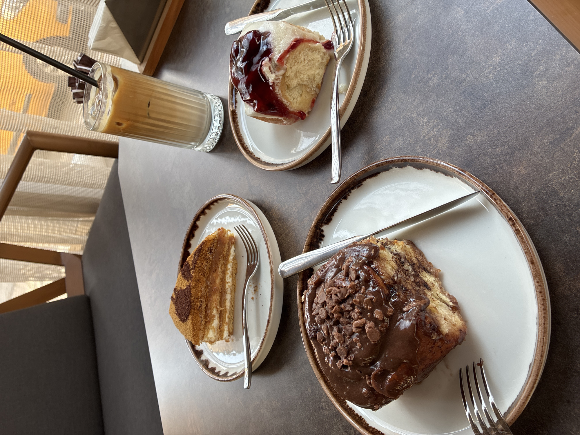 On a dark, textured table, three plates of desserts and a glass of iced coffee are arranged. In the foreground to the left, a large cinnamon roll slathered in chocolate frosting and topped with chocolate shavings sits on a white plate with a brown rim. A fork rests on the plate to the left of the roll. To the right of the foreground plate, a glass of iced coffee with a straw contains layers of coffee and cream, with ice cubes. Behind the glass, partially obscured by it, is a white napkin in a triangular holder. In the middle ground to the left, a slice of honey cake with visible layers of cake and cream, topped with a dusting of cocoa powder and brown sugar, is on a white plate with a brown rim. A fork lies on the plate. In the middle ground to the right, a sweet roll, likely a cinnamon roll, is topped with white icing and a generous dollop of dark red berry jam or compote. It is on a white plate with a brown rim, with a fork and knife to its right. In the background, a dark upholstered chair is visible, and behind it, sheer curtains are illuminated by natural light, suggesting an indoor setting, possibly a cafe. The overall scene conveys a sense of indulgence and relaxation. The style of the cafe and the desserts suggest a contemporary European influence, fitting for a city like Alanya, Turkey, which is known for its blend of Turkish and international cultures and its popular cafe scene, particularly along the coastline or within its bustling city center.