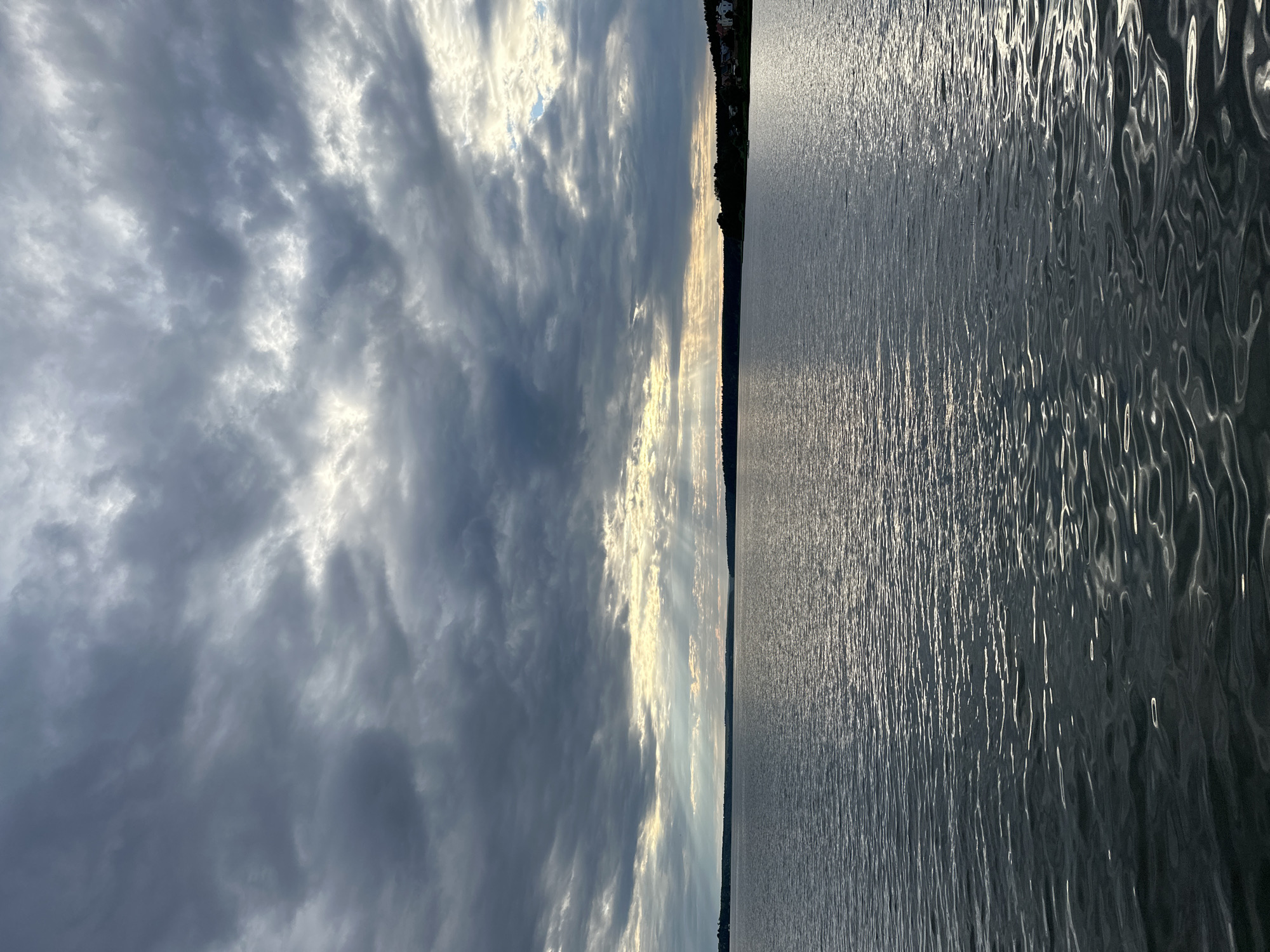 The image depicts a wide, calm body of water under a dramatic, cloudy sky. The clouds are a mix of dark grays and lighter blues, with shafts of sunlight breaking through in the distance. The water reflects the sky, creating shimmering patterns on its surface. A dark, tree-lined horizon separates the water from the sky, with a few scattered buildings visible on the right side of the horizon. The overall scene suggests an overcast day with intermittent sunlight. The location is identified as Votkinsk, Russia.