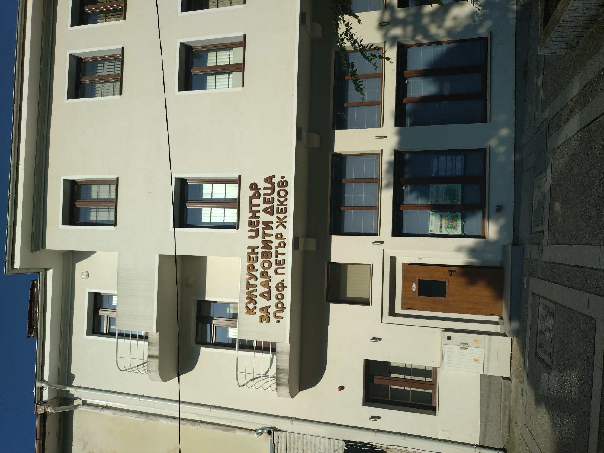 A multi-story building, identified as the "Cultural Center for Gifted Children 'Prof. Petar Zhekov'" by a prominent three-line brown sign in Cyrillic, stands under a clear blue sky in Stara Zagora, Bulgaria. The building has an off-white or light cream facade and a modernist architectural style.
The upper floors feature multiple rectangular windows with dark brown frames, many showing internal vertical blinds. The second floor includes two balconies with silver metal railings: a curved one on the left and a flat one to its right. Strong sunlight from the upper right casts distinct shadows across the building's facade, particularly from the balconies and the prominent horizontal band above the ground floor where the sign is located.
At ground level, a dark wooden entrance door with a small rectangular window is visible, situated to the right of a light-colored electrical utility box. A white drainpipe runs vertically down the left side of the building. The immediate foreground consists of a paved walkway with geometric patterns and a metal manhole cover. Tree branches with green leaves are visible on the far right, casting dappled shadows onto the lower windows of the building. No people are present in the scene.