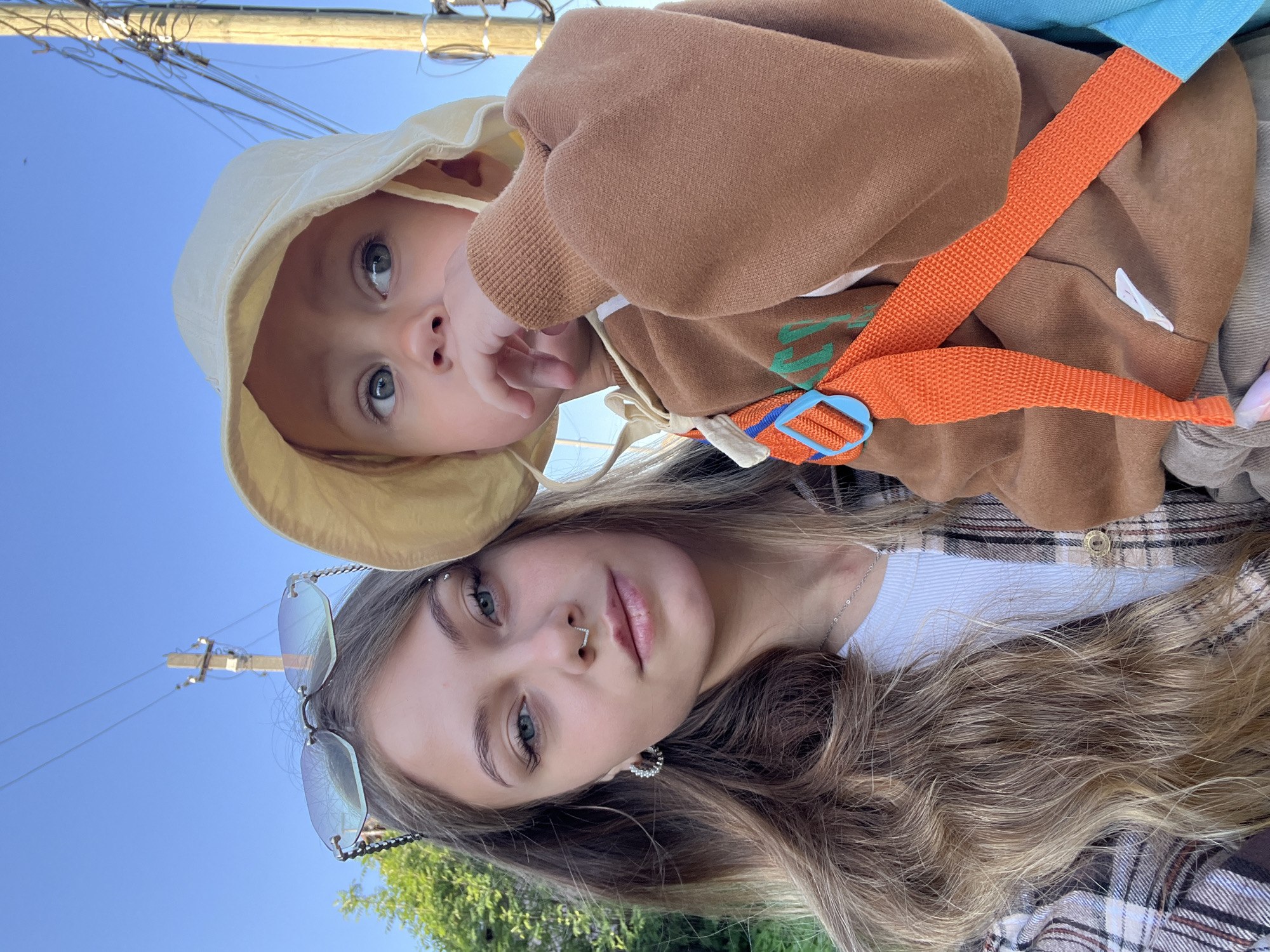 A woman and a baby are in the foreground. The woman has long, blonde hair and is wearing sunglasses perched on her head, a white tank top, and a brown and tan flannel shirt. She has an eyebrow piercing and a nose ring. The baby is wearing a beige sun hat and a brown sweatshirt with an orange backpack strap. The background is a bright blue sky with power lines and poles. There is greenery in the lower left corner. The photo is taken from a low angle, looking up at the subjects. The location appears to be outdoors, possibly a residential area, but the specific city of Фоки, Russia, cannot be verified from the image alone.