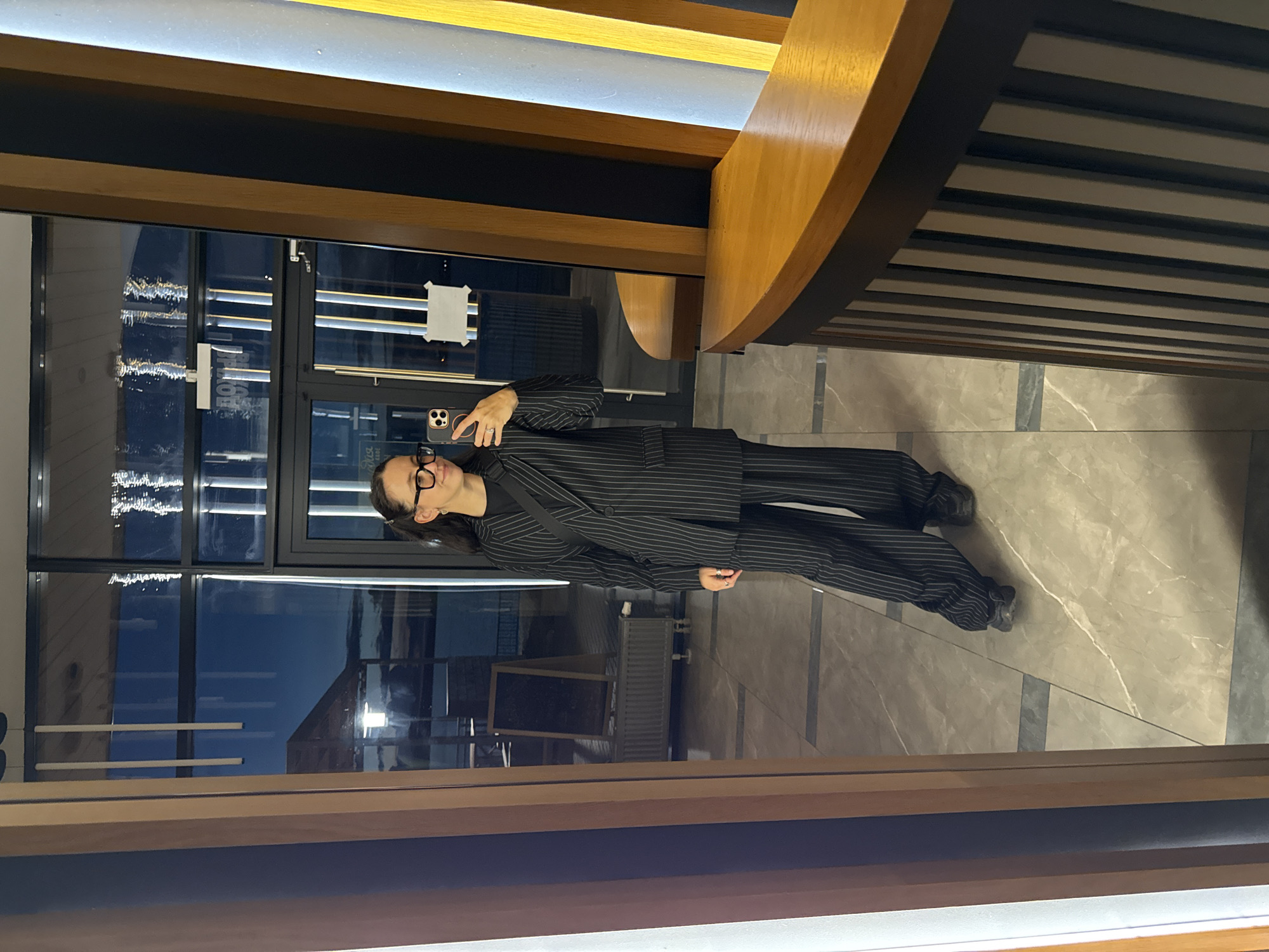 In Votkinsk, Russia, a woman stands in a hallway, taking a selfie in a large mirror. She is wearing a black pinstripe suit with wide-leg pants and a blazer. The woman has dark hair pulled back in a ponytail and is wearing glasses. She is holding a smartphone in her right hand, capturing her reflection. The background of the image shows large glass doors with a view of the outdoors, including some seating areas and a body of water. The interior features a marble-tiled floor and wooden accents.