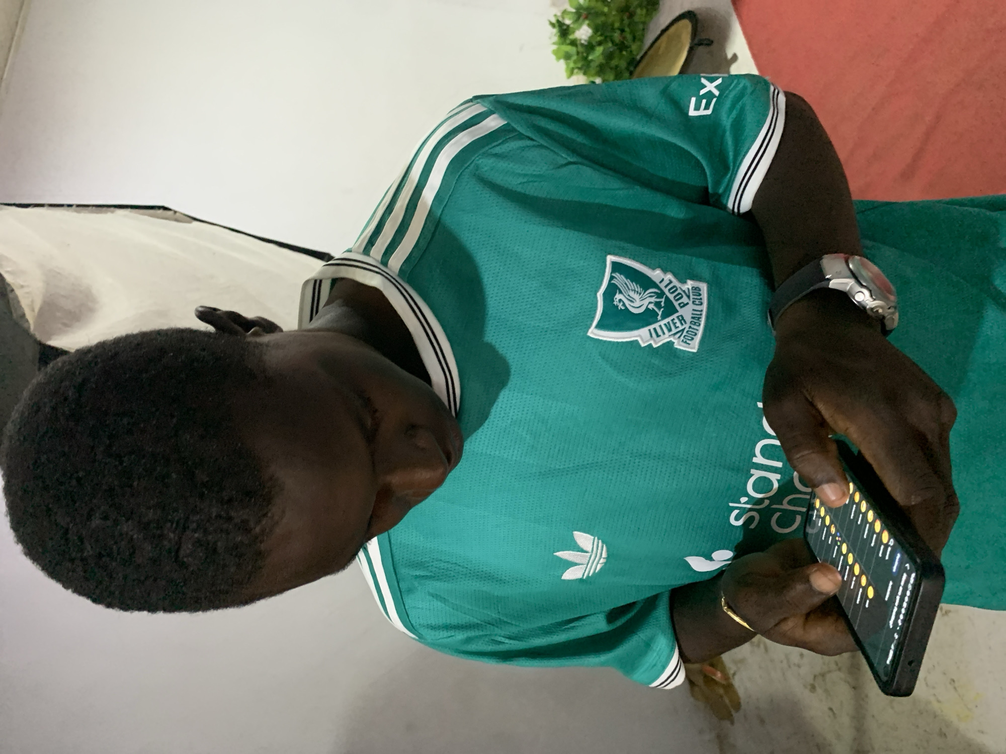 In the city of Jos, Nigeria, a young Black male is captured from the chest up, engrossed in his smartphone. He wears a vibrant green athletic jersey with white stripes on the shoulders and a crest featuring a Liver bird emblem. The jersey also displays a white Adidas logo and text on the chest that reads "Standard Chartered". The background is a plain, light-colored wall, with a white softbox light positioned behind him and to the right. To the far right, a cluster of green leaves is visible, and a red surface is partially in view at the bottom right of the frame. The man's hands are actively engaged with the smartphone, which displays a grid of yellow circles and lines, suggestive of a game or interactive application. He is wearing a silver wristwatch on his left wrist.