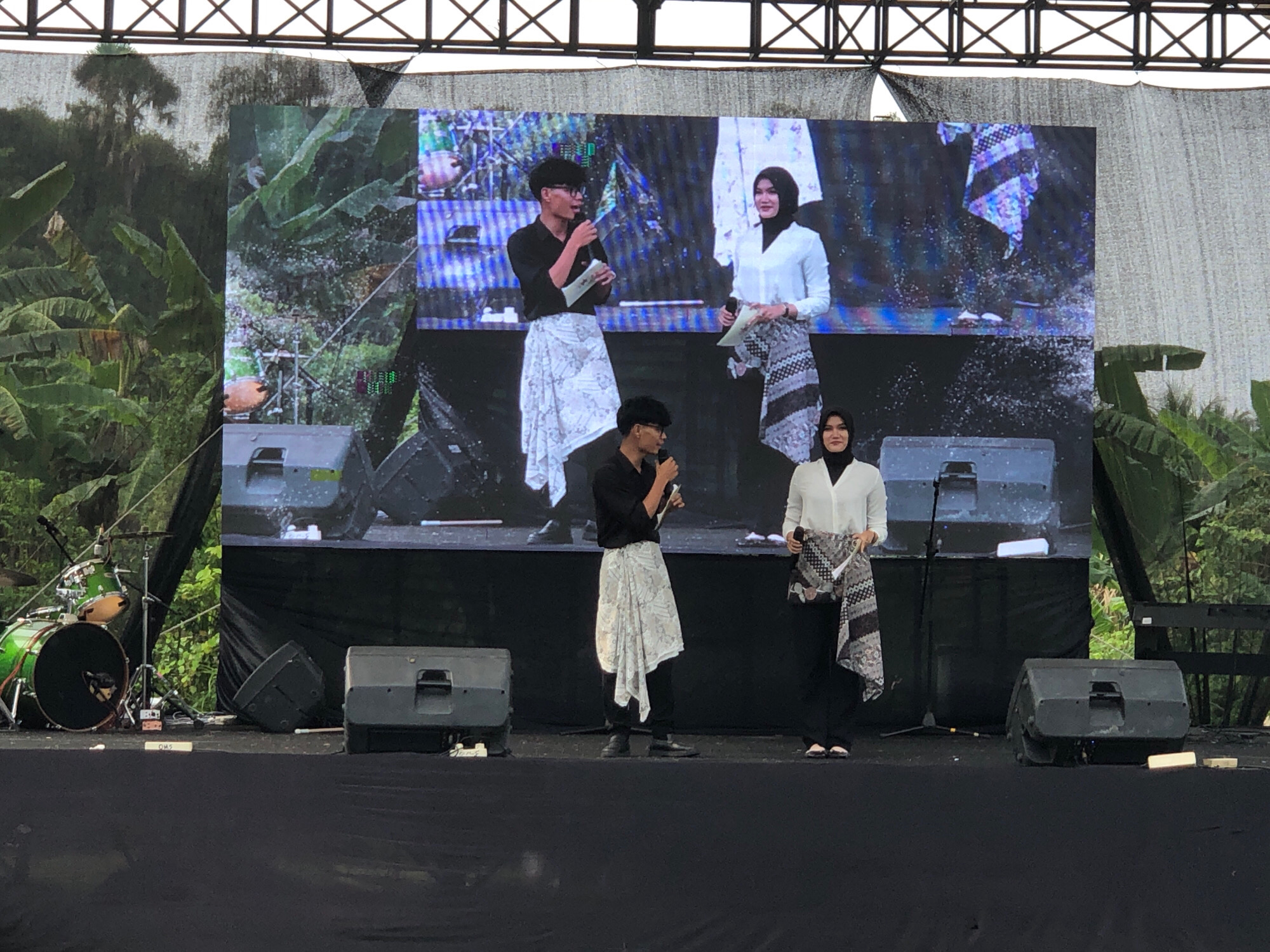 An outdoor stage event is taking place in Lhokseumawe, Indonesia, with a large LED screen as a backdrop. On the screen, two individuals are depicted, a man in a black shirt and white patterned skirt, and a woman in a white shirt and patterned skirt, both holding papers. In front of the screen, on the stage, four individuals are present. To the left, a man in a black shirt and white patterned skirt is speaking into a microphone. To his right, a woman in a white shirt and black pants, wearing a hijab, is holding a patterned fabric. Further to the right, another woman in a white shirt and black pants, also wearing a hijab, is looking forward. Behind them, stage equipment is visible, including speakers and a drum set to the far left. The overall environment suggests a cultural or promotional event.