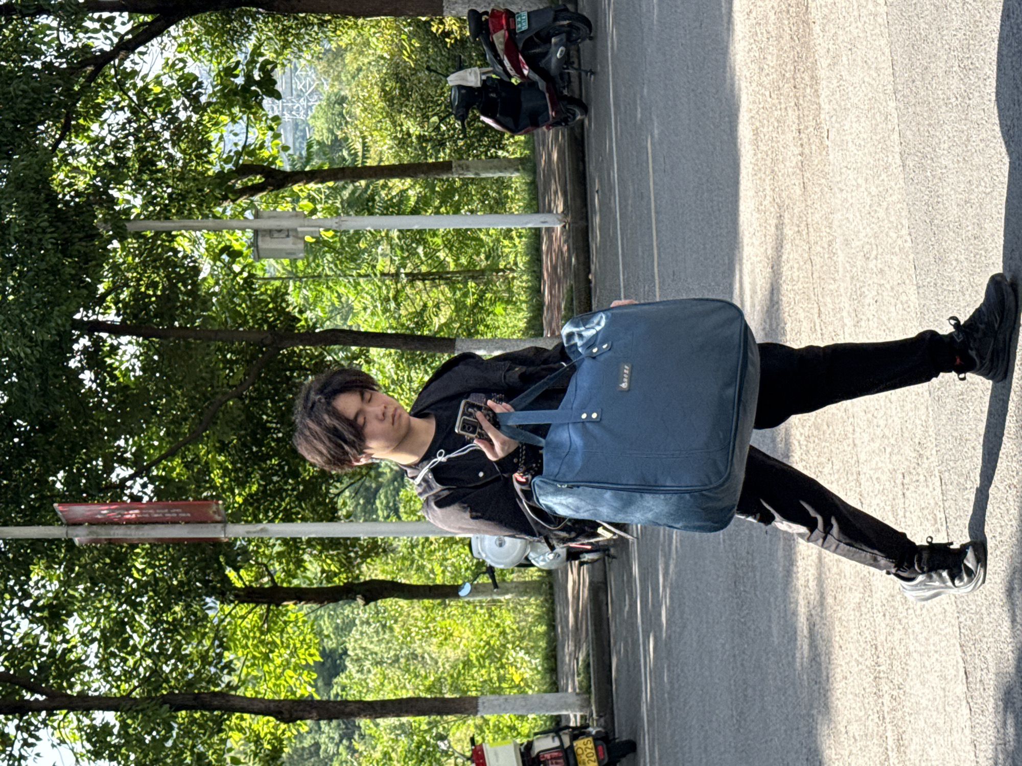 The image shows a person standing on a street in Jiangjin, China. The person is holding a mobile phone in their hand and carrying a blue bag. The person is standing beside a parked motorcycle, with other motorcycles parked behind it. The street is lined with trees. The environment appears to be outdoors during the daytime. The license plate is partially visible on one of the motorcycles.