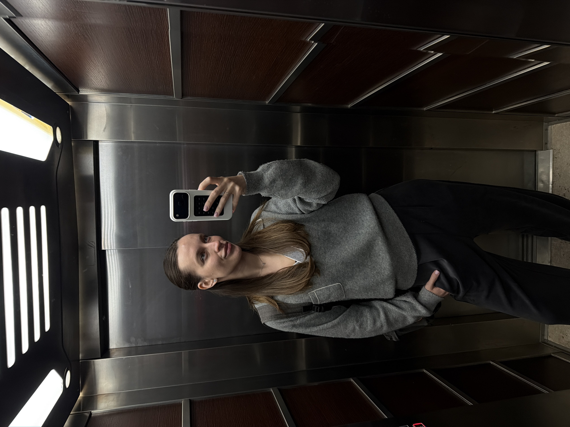 A woman is taking a selfie in an elevator. She is wearing a gray sweater and black pants. The elevator has stainless steel walls and a brown wood-paneled ceiling. The lighting is bright. The photo was taken in Budva, Montenegro.