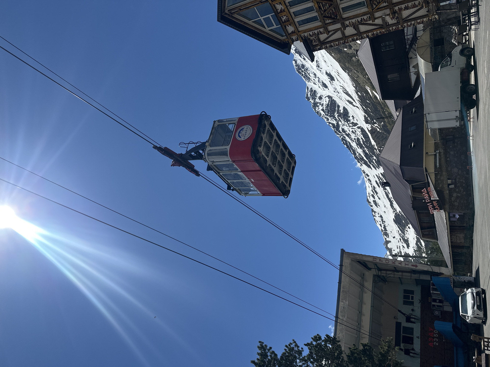A red and white cable car hangs suspended from cables against a clear blue sky, casting shadows on the ground. The cable car has a circular emblem on its side with text in Russian. Below the cable car, a snow-capped mountain range forms the backdrop. In the foreground, several buildings are visible. To the left, a concrete structure with a blue section contains the number "2350" and Cyrillic text. A white sedan with a license plate is parked in front of this building. To the right, a wooden-framed building with a dark roof is visible, and next to it, another building with a dark roof and illuminated windows. A white truck with its rear doors open is parked in front of this structure. The scene appears to be a mountain resort area in Terskol, Russia, likely a ski lift station. The sun is bright, creating lens flare in the upper left corner of the image.