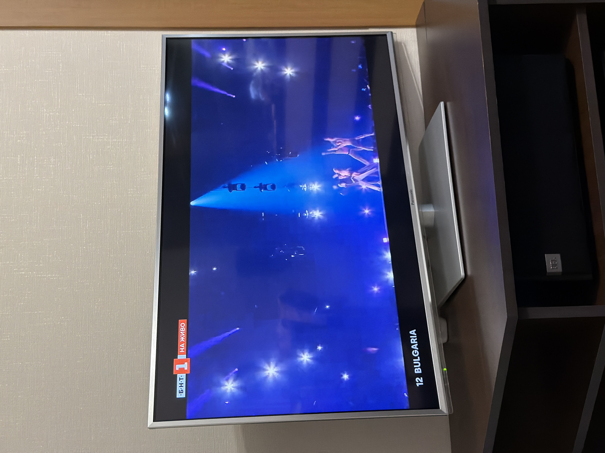 The image shows a television screen displaying a performance with blue lighting and performers, possibly a live broadcast as indicated by the "НА ЖИВО" (live) text. "12 BULGARIA" is visible on the screen, indicating a Bulgarian entry in a competition, possibly Eurovision. The television is mounted in a cabinet or shelving unit. The surroundings suggest an indoor setting. There is no information available in the image to confirm that it is in Burgas, Bulgaria.