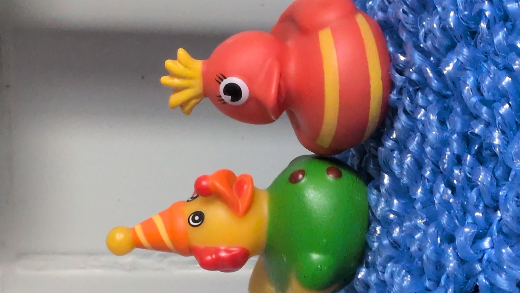 Two rubber ducks are positioned on a blue woven surface. The duck on the left is yellow with a green body, an orange and yellow striped party hat, and red clown cheeks. It has two brown spots on its chest. The duck on the right is red with yellow stripes and a yellow spiky headpiece. It has one eye with eyelashes. The background is a pale, textured wall. The location in the city of Rastenberg, Germany is not discernible from the image.