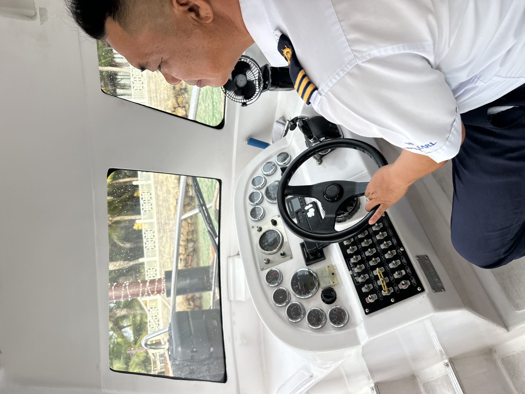 A male boat captain, seen in profile from the right side, pilots a white vessel. He wears a white short-sleeved uniform shirt featuring epaulets with three gold stripes and a gold anchor/ship's wheel emblem on the right shoulder, and dark blue trousers. Blue embroidery on his right sleeve cuff reads "WHITE PEARL." His right hand is positioned on the black steering wheel of the boat's helm station. The dashboard is white and equipped with a multi-gauge cluster, featuring approximately ten circular dials, and a rectangular panel below them containing at least twenty toggle switches. The text "HE DIEN 24V" is visible above this switch panel. A small black portable fan is mounted to the right of the captain. Through the front windows, which are speckled with water droplets, a coastal scene is visible. The boat is in light green, shallow water. On land, a retaining wall, numerous palm trees, tropical vegetation, and white decorative railings are present. A large reddish-brown cylindrical pillar stands amidst the foliage. A black case is visible on the boat's bow. The setting is a tropical coastal environment, consistent with Nha Trang Ward, Vietnam.