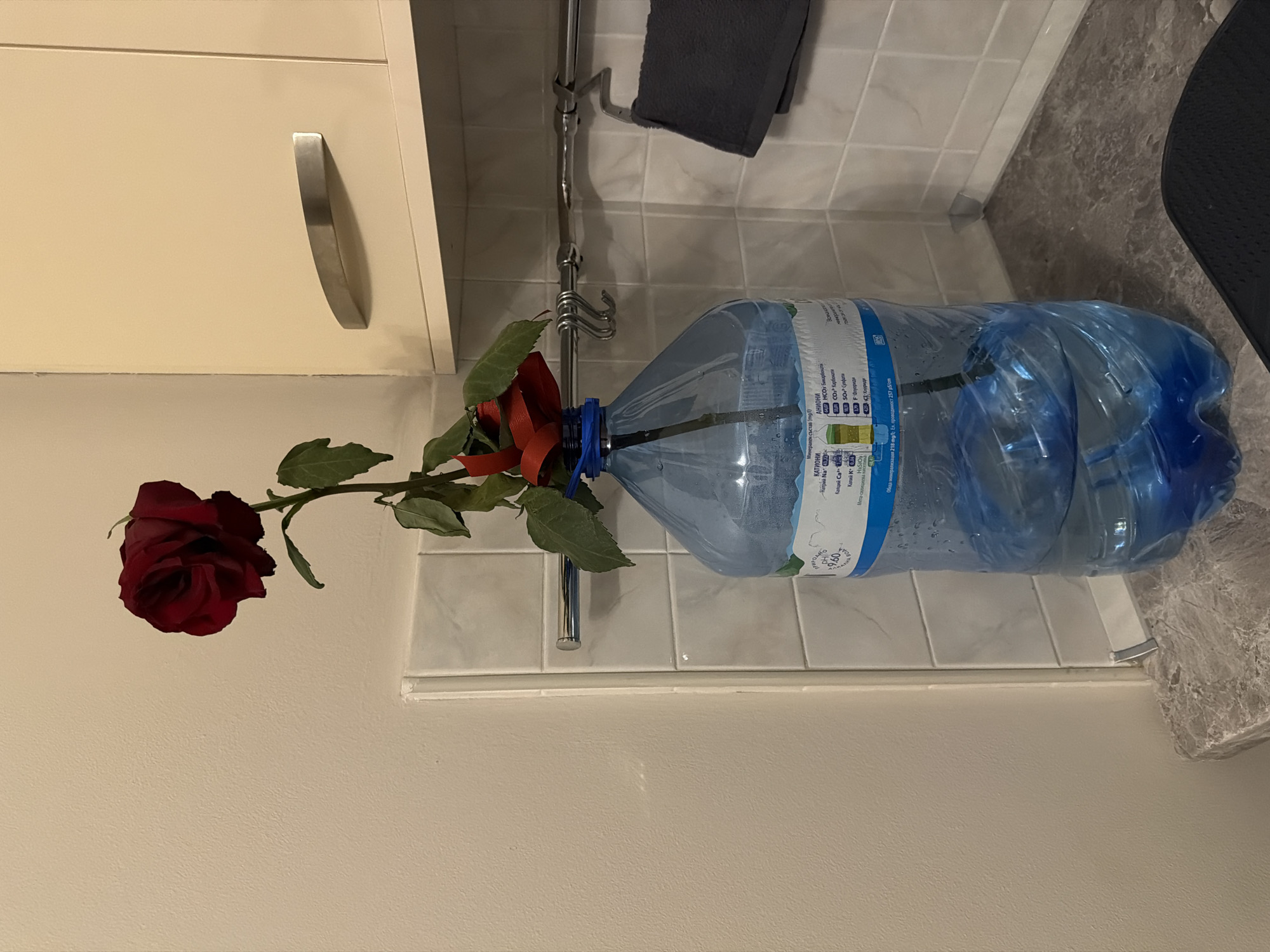 The image captures a domestic kitchen interior in Sofia, Bulgaria, identified by the Bulgarian text detailing mineral composition on the water bottle label. A large, approximately 5-liter, transparent blue plastic water bottle, filled with water, serves as a vase on a mottled grey countertop. A single deep red rose with green foliage is centrally placed within the bottle, its stem submerged. A smaller red element, possibly a ribbon or petal, is visible near the bottle's neck. The background features a light beige painted wall on the left and white square tiles on the right. An overhead cream-colored cabinet with a silver curved handle is positioned above the tiled wall on the right. A horizontal silver metal rod is mounted on the tiled wall, equipped with multiple silver S-hooks on the left and a dark grey hand towel draped over a separate hanger on the right. A section of a black, textured mat is visible on the countertop in the lower right foreground. No individuals are present.