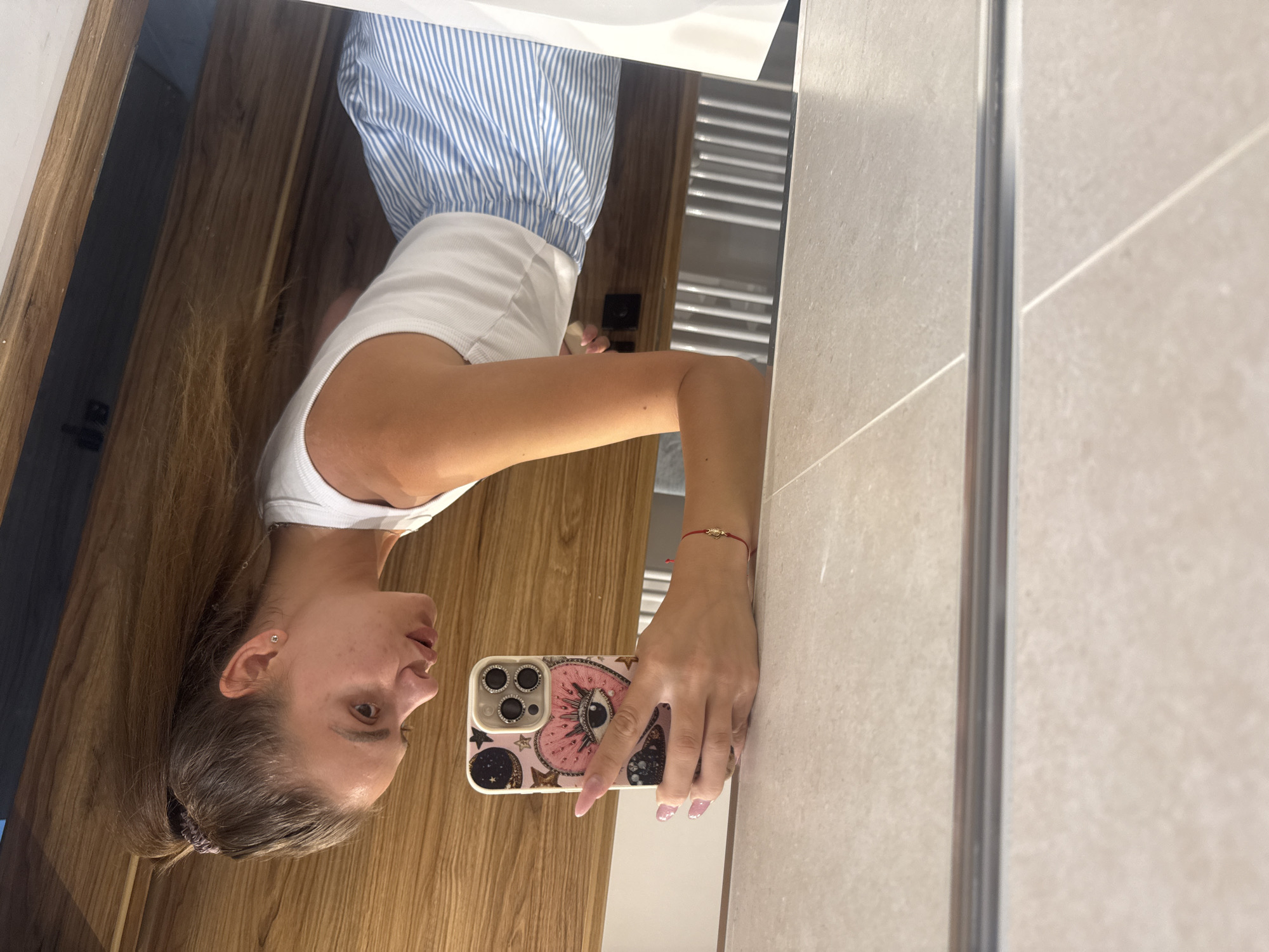 An indoor mirror selfie captures a young woman standing before a large rectangular mirror. The woman is facing slightly to her left, holding a smartphone with her right hand to take the photo. She has long brown hair pulled back, revealing a simple stud earring in her left ear, and her expression is neutral.
She is wearing a white sleeveless top and blue and white striped bottoms, possibly shorts or a skirt. A delicate red string bracelet with a small, gold-colored charm is visible on her right wrist. Her fingernails are manicured with a light pink polish. The smartphone case is distinctive, featuring a large eye design with eyelashes and several small stars on a pink and gold background.
The background reflected in the mirror suggests a modern interior, possibly a bathroom or dressing area. To the woman's left, a warm-toned wooden door or wall panel is reflected. To her right, a light-colored, possibly beige or grey, tiled wall with grout lines is visible. A white edge, likely part of a sink or countertop, runs along the top right of the reflection. Further back in the reflection, a white ladder-style radiator can be seen on the tiled wall. The lighting appears to be ambient indoor light, and no visible text is discernible.