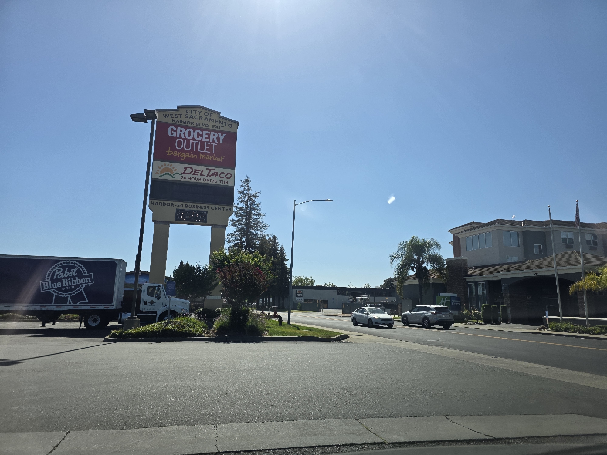 This media file captures a sunny daytime scene in West Sacramento, USA. The image showcases a commercial area with a prominent signpost advertising "CITY OF WEST SACRAMENTO HARBOR BLVD. EXIT" with "GROCERY OUTLET bargain market" and "DELTACO 24 HOUR DRIVE-THRU" listed below. A Pabst Blue Ribbon beer truck is parked on the left side of the frame.
In the background, a Holiday Inn Express hotel is visible, with cars driving on the road in front of it. The sky is clear and blue, indicating fair weather. The presence of palm trees suggests a warm climate. The overall impression is a typical suburban commercial landscape on a bright, clear day. No people are clearly visible in the image, but the scene implies a busy retail and hospitality environment. The "HARBOR-50 BUSINESS CENTER" is also mentioned on the signpost.