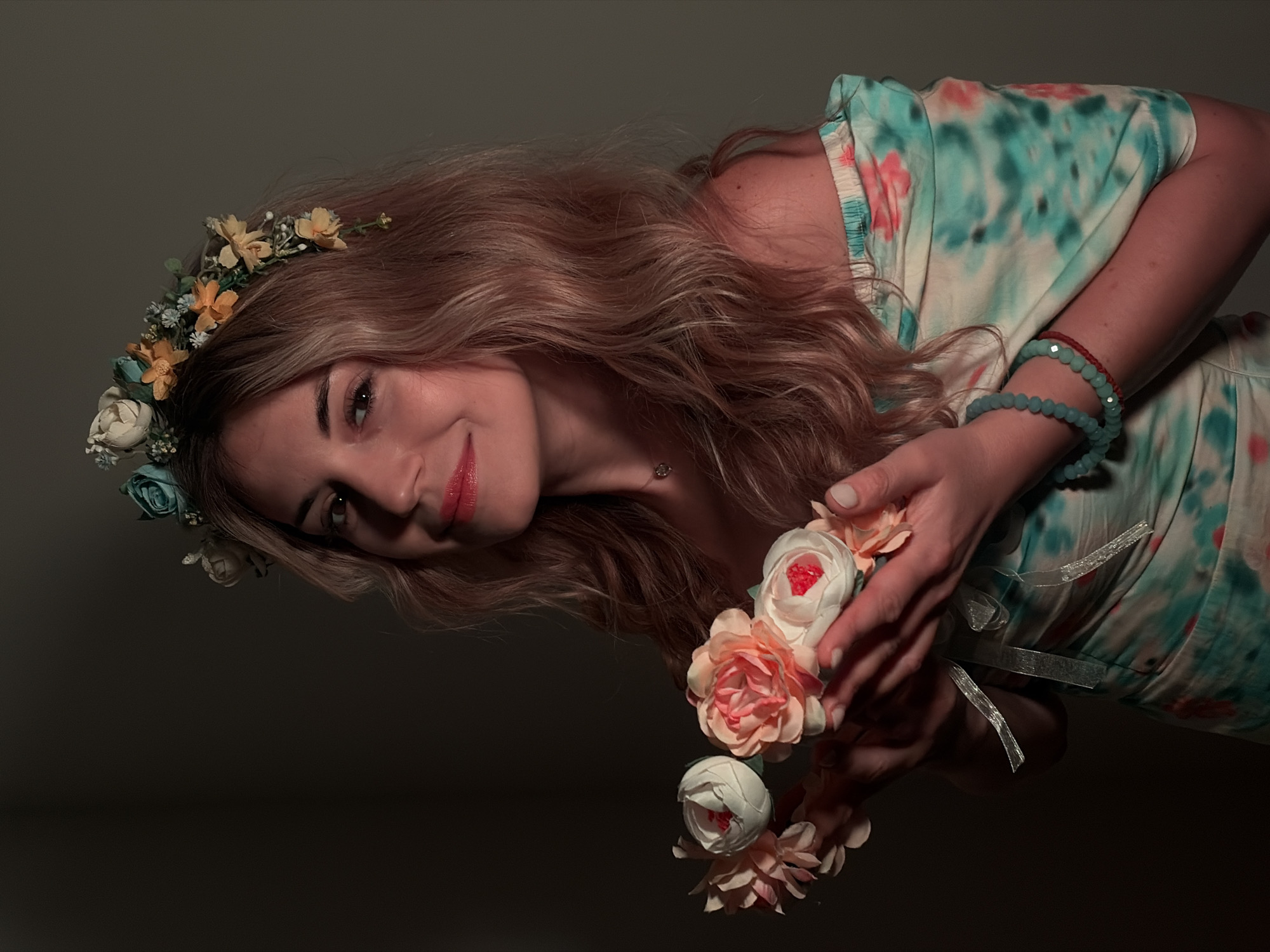 A woman with long, wavy blonde hair wearing a flower crown is holding a bouquet of flowers. She is wearing a dress with a blue and pink floral pattern and several beaded bracelets on her wrist. The background is dark, and the image is likely taken indoors. There is no information available about this image being taken in Budva, Montenegro.