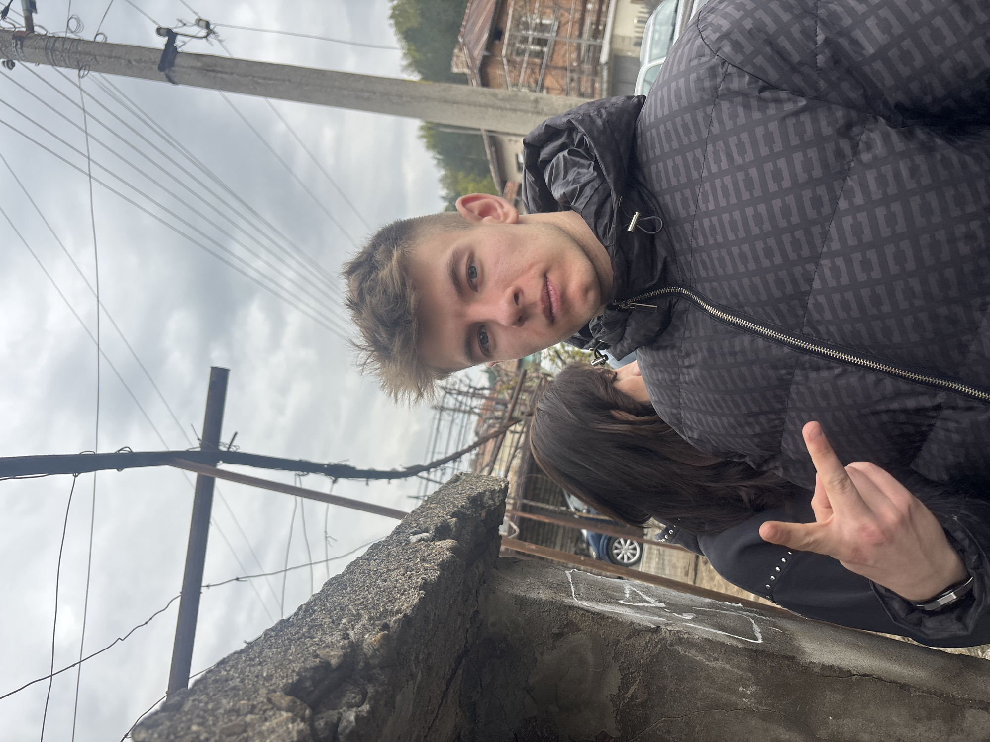 A medium shot, taken from a low angle, shows a light-skinned young man in a dark, patterned jacket, looking directly at the camera. He is standing next to a woman with dark hair, whose face is partially obscured, and who is making a hand gesture. The background includes a weathered concrete wall, a metal structure with wires, an electrical pole with hanging wires, buildings, trees, and a cloudy sky. The location is likely in Bratsigovo, Bulgaria, based on visual elements common to urban areas in that region.