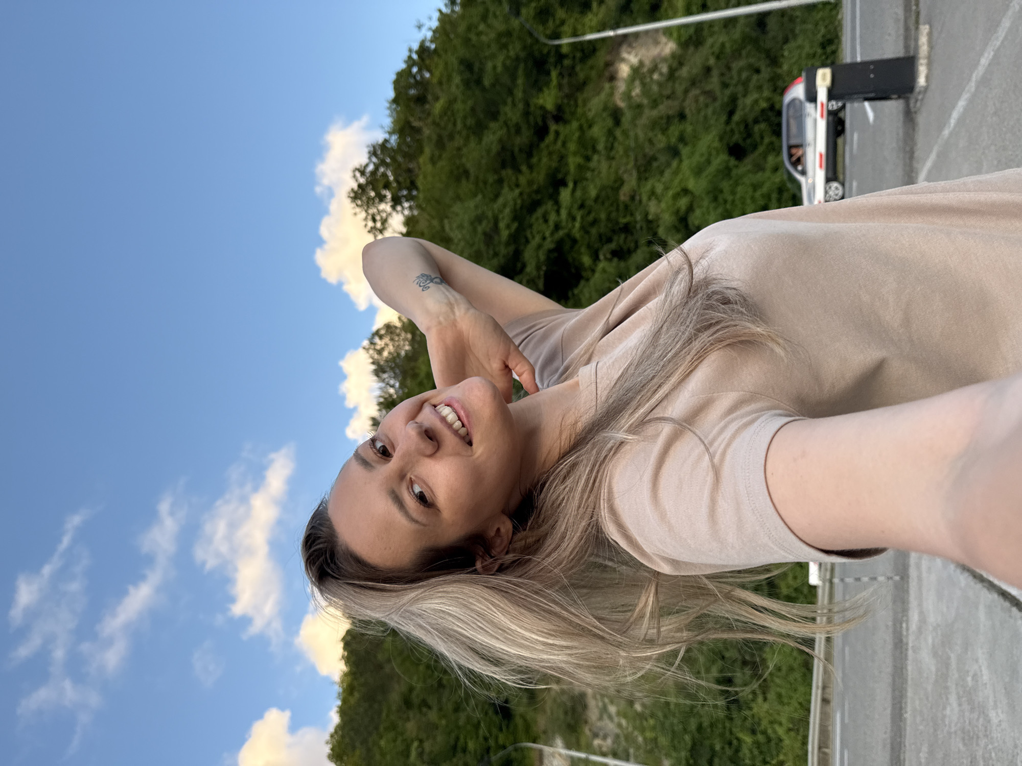 In Nebug, Russia, a young Caucasian woman with blonde hair, wearing a beige t-shirt, poses for a selfie. She is outdoors, with a lush green, tree-covered hillside forming the background. The sky above is blue with scattered white clouds. To the right, a metal barrier and a street lamp are visible, and a silver car is parked in the distance. Her left arm is raised behind her head, and she has a tattoo on her forearm. She is smiling directly at the camera.