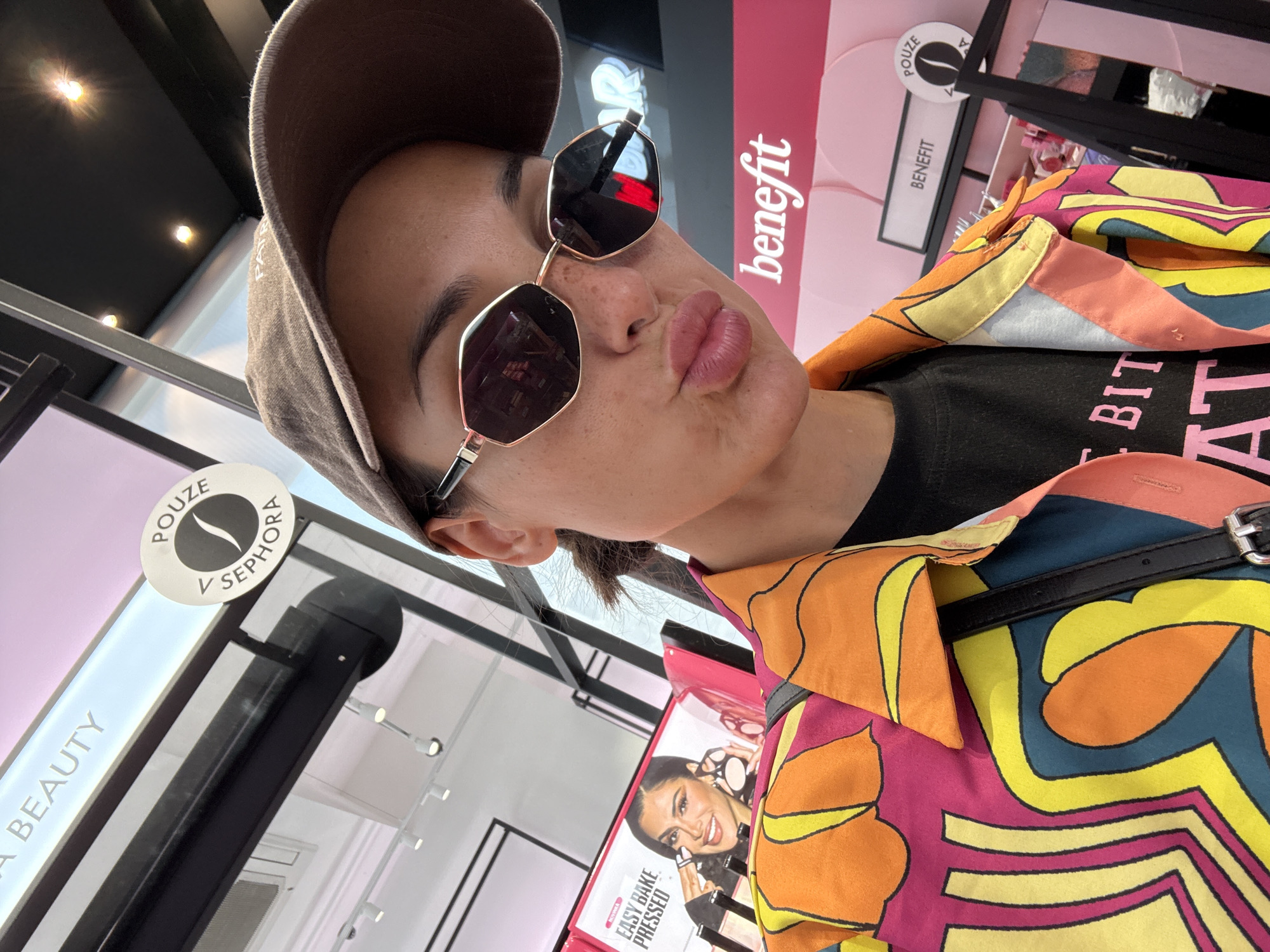 A woman wearing a brown baseball cap and sunglasses poses for a selfie in a brightly lit beauty store. She is wearing a vibrant, multi-colored shirt with a psychedelic pattern. Behind her, a display for the beauty brand Benefit is visible, with pink circular accents. A sign for Sephora is also present. The overall scene suggests a shopping environment within a commercial district.