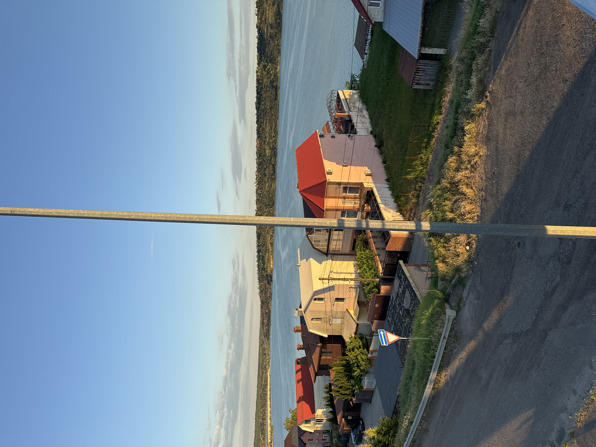 The image shows a waterside view of a residential area in Altestove, Ukraine, bathed in the warm light of what appears to be late afternoon or early evening. A wide body of water, likely a river or a bay, dominates the background. Across the water, a distant treeline and hills form the horizon, with some structures visible on the crest of the hills. In the mid-ground, several houses with pitched roofs, varying in color from pink to white and red, are nestled together. Some of the houses have multiple stories and are adorned with architectural details. A prominent light-colored metal pole, likely a lamppost or utility pole, runs vertically through the foreground and center of the image, casting a long shadow across a paved area in the lower left.
The foreground consists of an asphalted area, possibly a road or a driveway, showing signs of wear. Adjacent to this paved area is a patch of dry, sun-baked grass and weeds. A few trees and bushes are visible scattered amongst the houses and along the shoreline. A blue and white triangular traffic sign indicating a yield or give way is present near the road. Several small structures, possibly sheds or garages, are also visible among the residential buildings. The sky is a clear, pale blue with a few wispy clouds scattered near the horizon. No people are visible in the image.