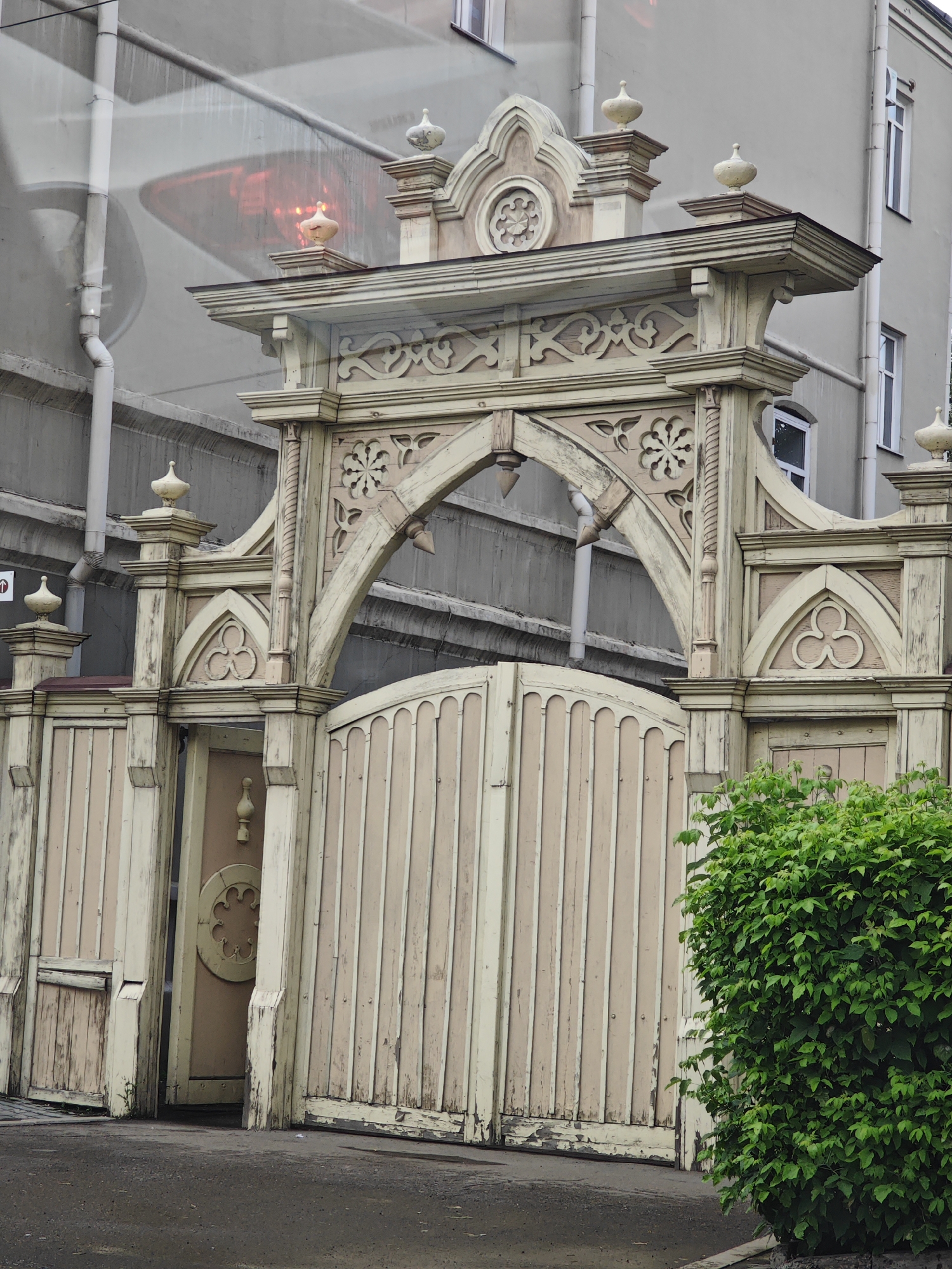 The image shows a decorative wooden gate in Krasnoyarsk, Russia, featuring intricate carvings and a faded pastel color scheme. The gate is adorned with ornate floral motifs and geometric patterns, with a central archway supported by decorative pillars. The main gate appears to be made of vertical wooden planks, and a smaller pedestrian gate is visible to the left. A lush green bush stands to the right of the gate. The background consists of a plain, light-colored building with windows and a white downspout. The overall impression is one of historical architecture and a touch of rustic charm.