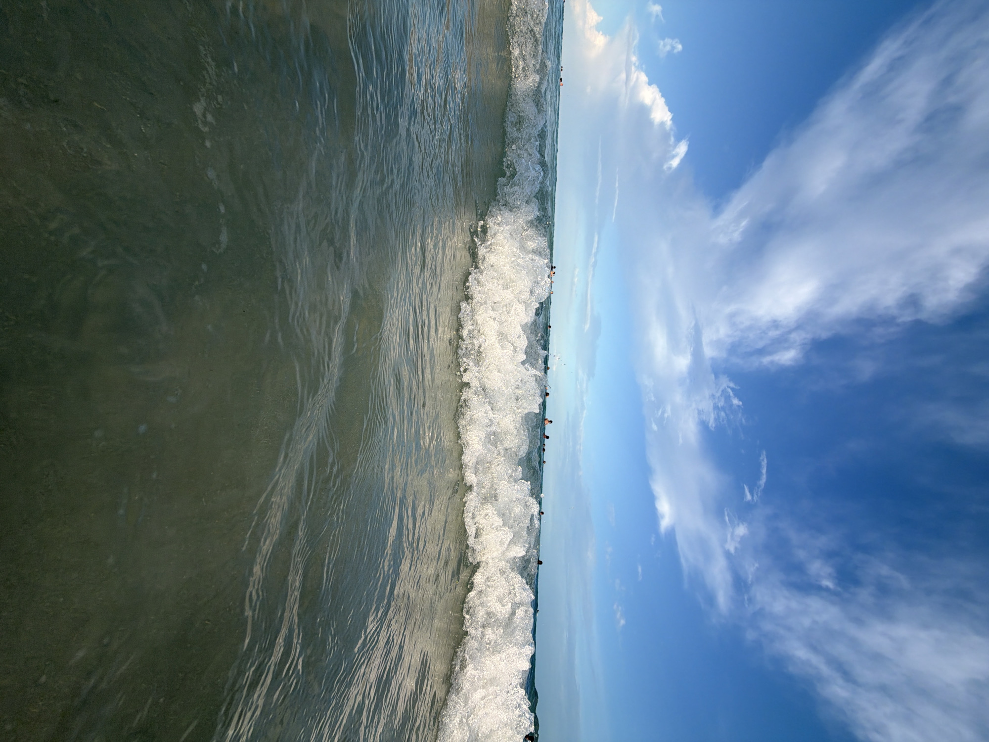 The image shows a low-angle shot of a breaking wave in the ocean. The water is clear with a greenish-blue hue, and the wave is white with foam. In the background, the sky is a bright blue with scattered white clouds. Several people are visible in the water in the distance, some appearing to be swimming or wading. The scene suggests a beach environment, likely in a tropical location given the clear water and bright sky. The location is identified as Ko Samui District, Thailand.