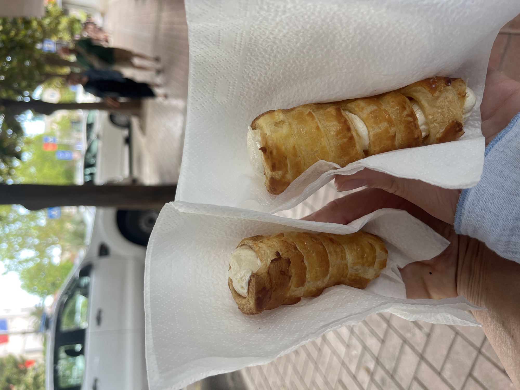 The photograph shows two cream horns, a type of pastry, held in hand with paper towels. The cream horns are golden-brown with a layered, flaky appearance, and filled with a white cream. A person's hand and part of their arm are visible, wearing a light blue top. The background includes a blurred street scene with a white car, trees, and other people walking. The setting appears to be outdoors, likely in a public area in Budva, Montenegro. The photograph is taken at close range, focusing on the cream horns. The environment is characteristic of a street scene.
