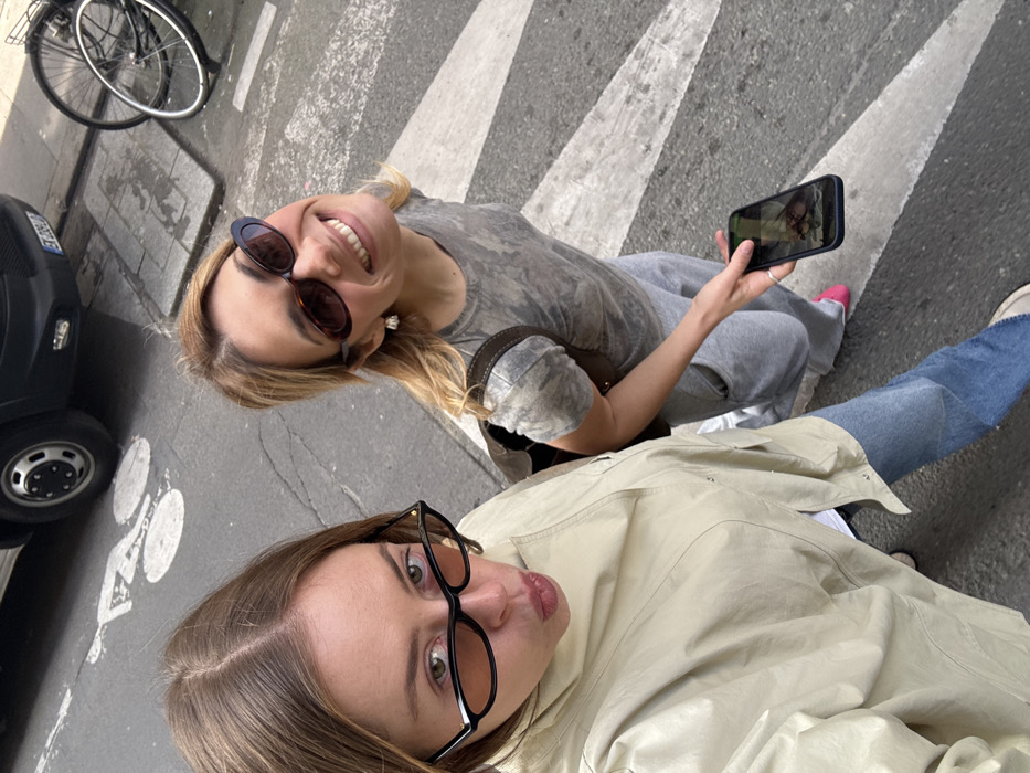Two women are standing on an asphalt street featuring white pedestrian crossing stripes and a white bicycle lane symbol. The woman on the left, with brown hair parted centrally, wears black oval sunglasses and a light beige collared shirt over blue jeans, with her lips pursed towards the camera. The woman on the right, with blonde hair parted to the side, wears dark brown oval sunglasses, a grey patterned t-shirt, and light grey sweatpants with visible pink footwear. She smiles broadly towards the camera, holding a dark smartphone vertically in her right hand with the screen displaying an image, and has a dark brown shoulder bag on her left shoulder. In the background, a dark-colored van's front wheel and lower bumper are partially visible, with a license plate fragment showing "EE". Two bicycles lean against an unseen object on a curb to the right. The scene is located in the city of Unknown, Unknown.