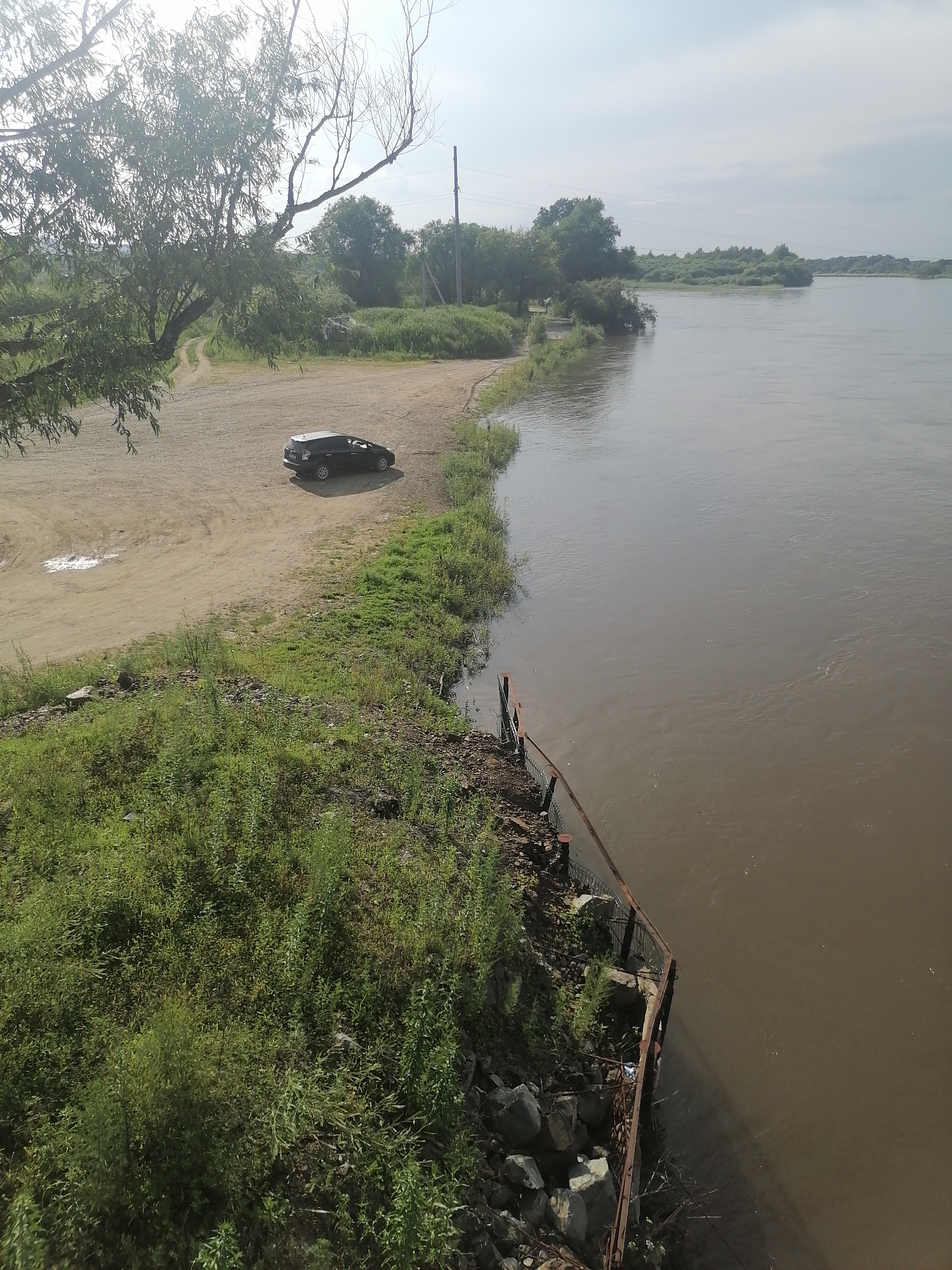 The image captures a wide, muddy river flowing through a natural landscape on a partly cloudy day. On the left bank, a dirt parking area is visible, with a black hatchback car parked. Adjacent to the parking area is a dense green area with various types of vegetation, transitioning to a rocky embankment with a rusty metal railing bordering the river. In the background, trees and foliage line the opposite bank, with a few power lines extending across the scene. The overall impression is one of a rural or undeveloped area, possibly on the outskirts of Novopokrovka, Russia, given the natural setting and the presence of a vehicle suggesting human activity.