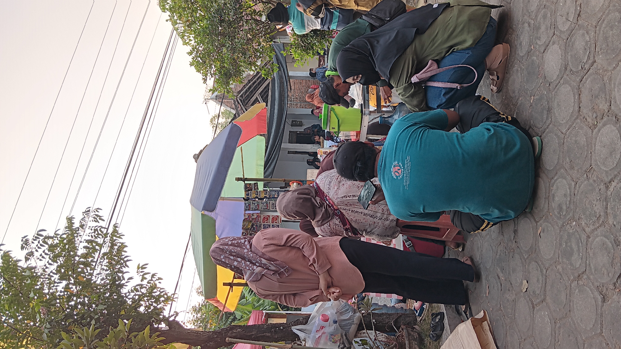 This image depicts an outdoor gathering in Cerme, Indonesia, likely a street food stall or community event. Several people are present, some standing and others seated on the ground or low stools.
In the foreground, a person wearing a teal t-shirt and dark pants is sitting on the ground, facing away from the camera. To their right, another person in a green jacket and blue jeans is crouched down. They are both looking towards a table laden with food and drinks. A striped umbrella, with panels of yellow, blue, green, and red, is set up over the stall area.
Behind the seated figures, a woman in a dusty rose shirt and black pants stands with her hands clasped behind her back, looking towards the stall. Next to her, another woman in a patterned top and hijab is seated, holding a phone.
In the background, more people are visible around the stall and in the vicinity of a building. The environment appears to be a paved area with trees and power lines overhead, characteristic of a semi-urban or rural setting in Indonesia. The overall atmosphere seems casual and communal, suggesting a daytime activity or evening gathering.