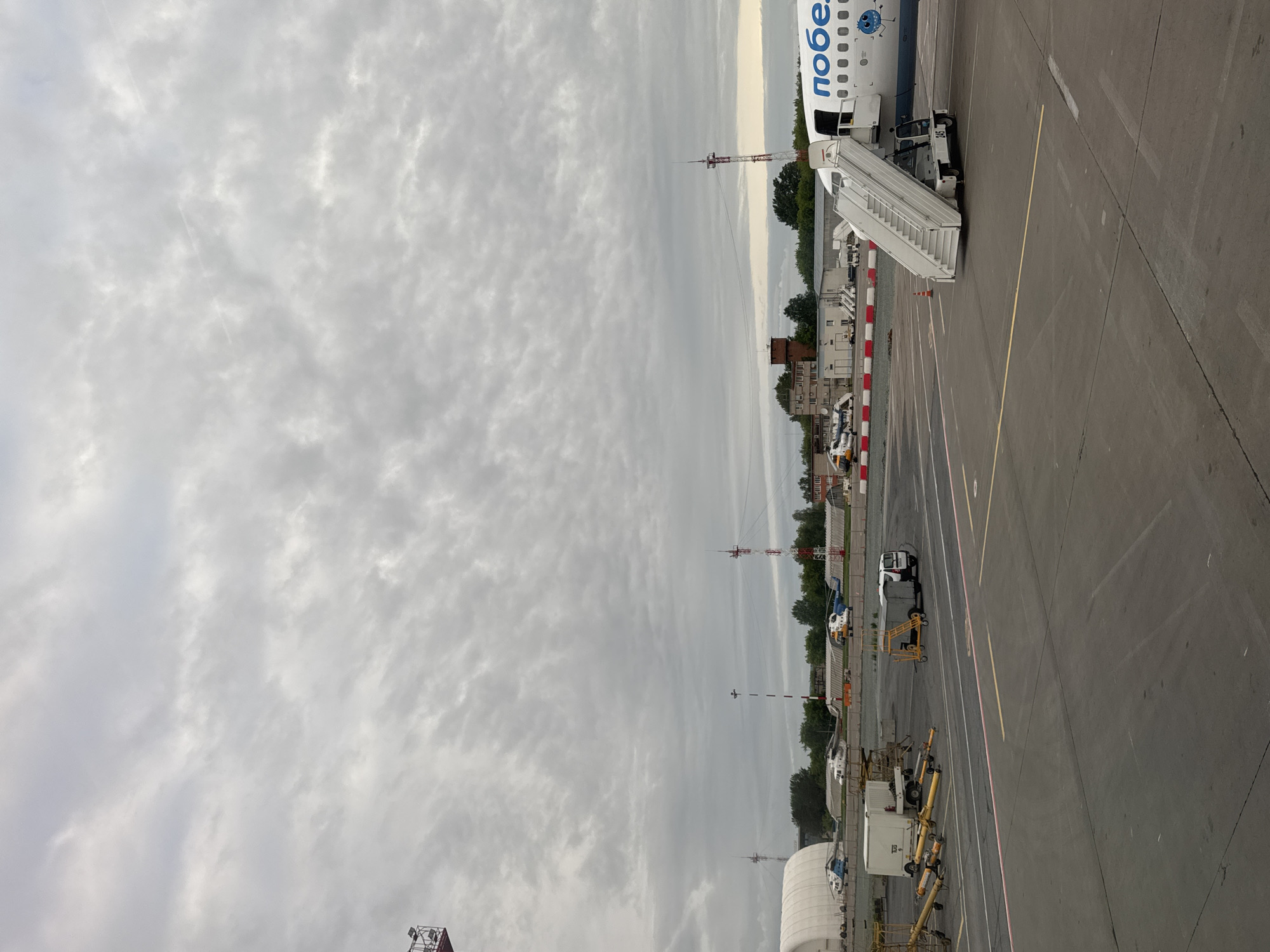 The image shows an airport apron under a cloudy sky. An airplane with "Победа" (Pobeda) written on its side is parked, with a set of stairs attached. Various ground vehicles and equipment are visible on the apron, including a small truck and several other utility vehicles. In the background, there are buildings and trees. The location is identified as Башкултаево, Russia.