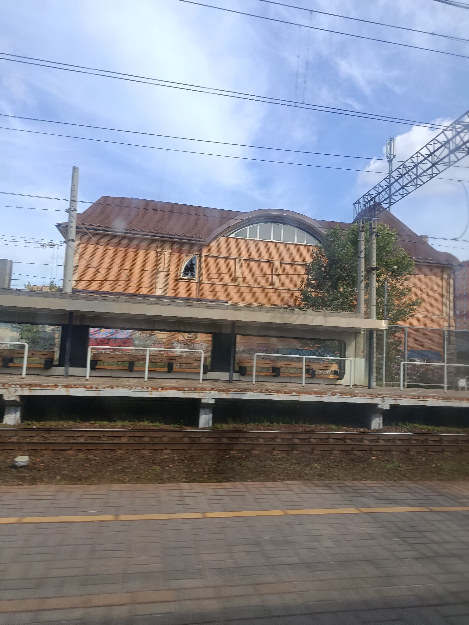 This is a daytime, eye-level, medium-shot photograph taken from a moving train. The scene captures a train station platform and building, likely in Pavlovsky Posad, Russia, given the context.
The train tracks are prominently in the foreground, with ballast and some sparse vegetation visible between them. A paved platform with a yellow tactile paving line runs parallel to the tracks. Behind the platform, a covered waiting area with wooden benches and white railings is visible. The back wall of this covered area appears to be covered in graffiti.
The main subject is a multi-story brick building behind the platform. The building has a red-tiled, pitched roof with a large arched window at the front. The brickwork is orange-red, and there are visible brick patterns on the facade. A small, broken window is visible on the side of the building. A tree with green foliage grows to the right of the building. Overhead, power lines and a section of a metal lattice structure, likely for supporting these lines or communications equipment, are visible against a blue sky with scattered clouds. To the far right, a portion of a modern apartment building is visible. The overall impression is that of a functional, somewhat aged, urban train station environment.