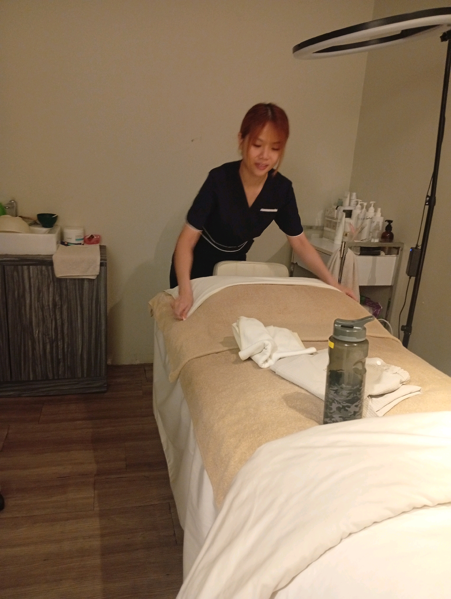 In a well-lit room, likely a spa or wellness center, a woman with reddish-brown hair, wearing a dark blue uniform, is seen preparing a massage bed. She is leaning over the bed, her hands are on the tan-colored sheet, appearing to adjust or smooth it. The massage bed is covered with a tan sheet and white linens. On the bed, a dark grey water bottle with a black lid is placed near the head of the bed, alongside folded white towels. In the background, to the left of the woman, is a grey cabinet with a white sink on top, with a small green bowl and other items. To the right of the woman, a tiered trolley holds various bottles and containers, suggesting spa products. A ring light on a stand is positioned above and behind the woman, providing bright illumination. The room has a neutral-colored wall and a wooden floor. The overall scene depicts a professional setting where a client is about to receive a service. The location is specified as Selayang Municipal Council, Malaysia, which implies this is a commercial establishment within that administrative area.