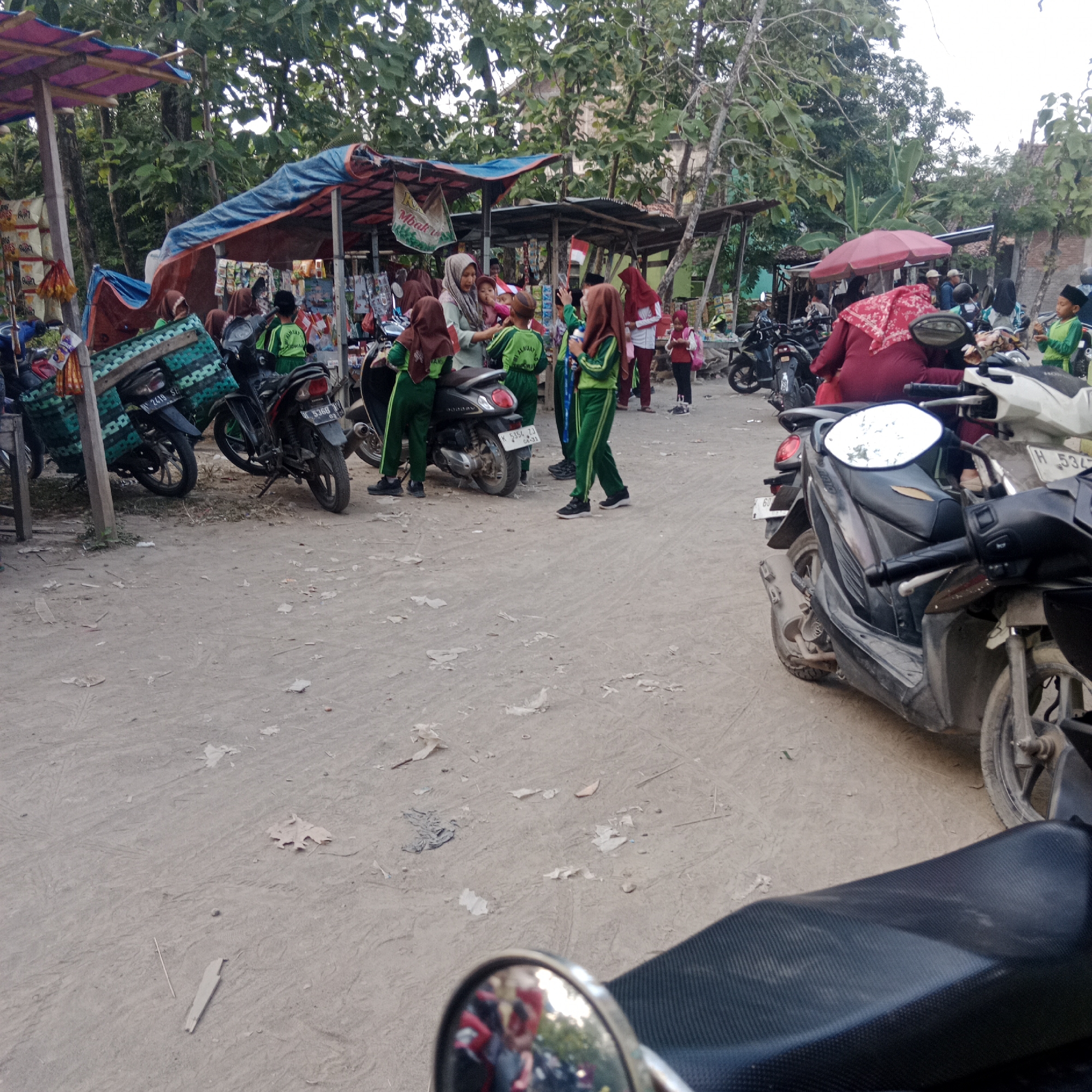 The image captures a scene in Tegowanu Kulon, Indonesia, likely near a school or market. The foreground features the seat and handlebars of a motorcycle. In the midground, several other motorcycles are parked on a dirt ground. A group of children, some in green and brown school uniforms with headscarves, are gathered around. Some are interacting, while others appear to be waiting. Behind them, makeshift stalls with colorful awnings, possibly selling goods, are set up. Trees and foliage line the background, suggesting an outdoor, possibly semi-rural setting. The overall impression is one of a busy gathering place, perhaps a collection point for students or a local market in operation.