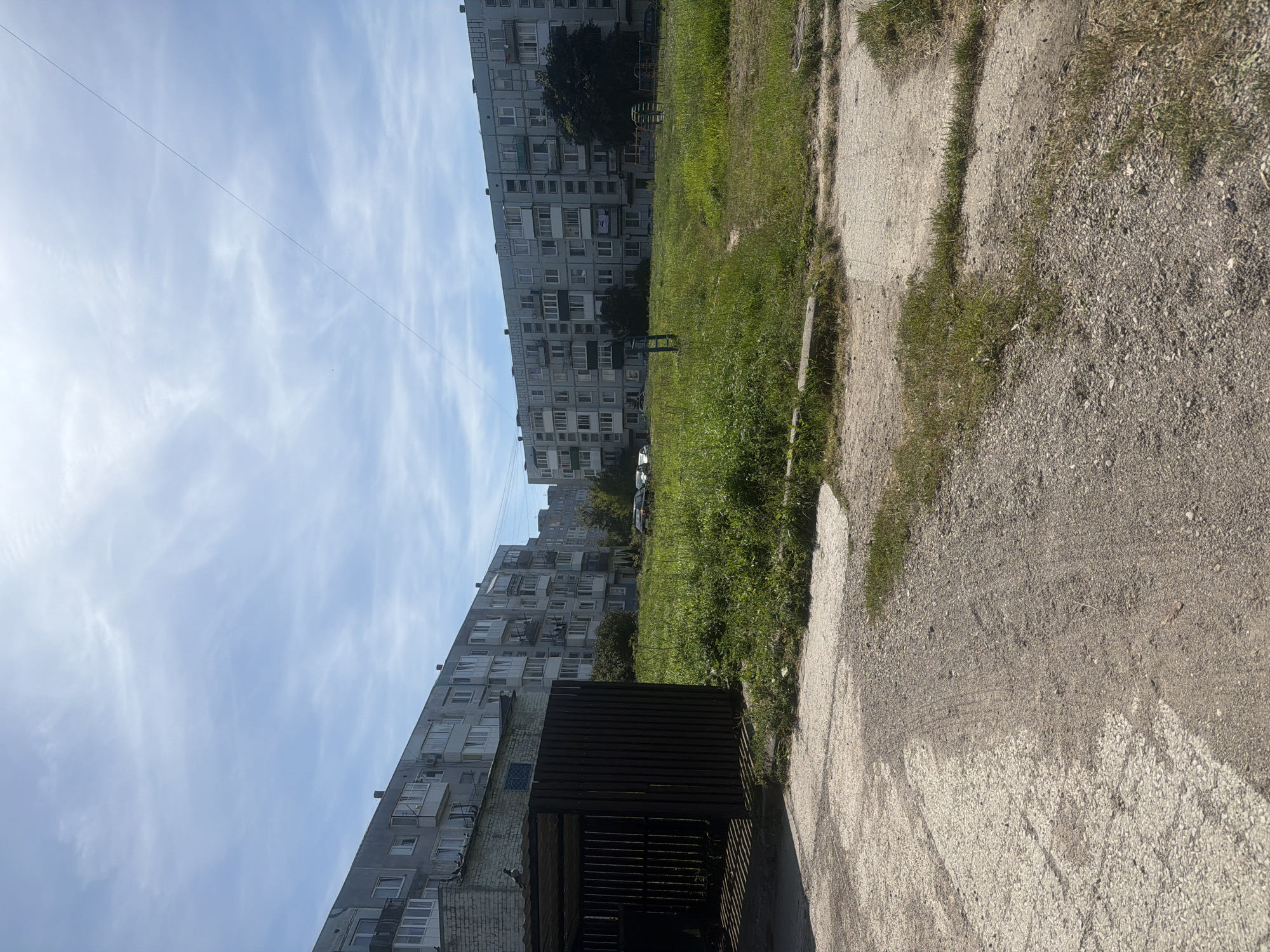An outdoor, eye-level shot, taken from a low-angle, captures a residential courtyard in Baltiysk, Russia on a clear, sunny day. The foreground is dominated by a cracked, dirty concrete path with some patches of sparse grass growing through it. To the left, a dark wooden, open-sided structure, possibly a carport or shelter, is visible. Behind this structure and to the left is a portion of a two-story brick building with a light-colored wall.
The midground features a large, unkempt grassy area with a few scattered patches of concrete or asphalt. In this area, there is a blue metal pole and some playground equipment, including a colorful climbing structure. Several cars are parked on the grass or on the edge of the grassy area.
The background is comprised of several large, multi-story apartment buildings, characteristic of Soviet-era architecture. These buildings are gray and appear to be in a state of moderate wear. They are lined with numerous windows and balconies, some of which have air conditioning units or visible clutter. The sky above is a bright blue with wispy white clouds and a faint power line stretches across it from the right side of the frame towards the left.