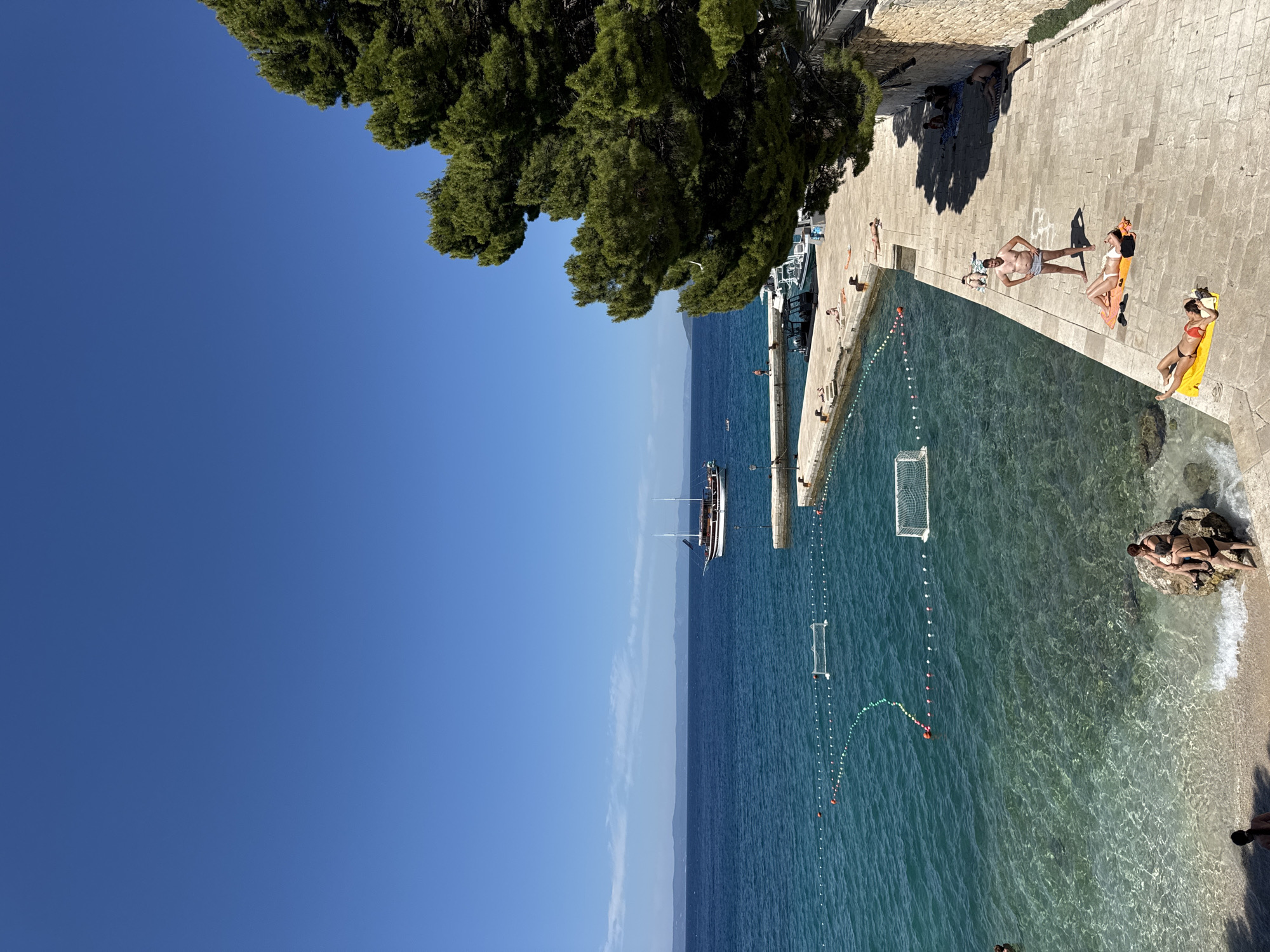 The image depicts a sunny day at a beach and promenade in Makarska, Croatia. The clear blue sky stretches over a calm, turquoise sea. In the water, there are two floating soccer goals, suggesting an area designated for water sports. A classic sailing boat with two masts is anchored in the distance.
The foreground shows a stone-paved promenade lining the edge of the water. Several people are relaxing on the promenade. One man stands shirtless, wearing swim trunks, while two women lie on towels, sunbathing. Further down the promenade, more individuals are seated or lying on towels, enjoying the sun.
To the right, a large, mature pine tree with lush green foliage overhangs the scene, casting shadows on the promenade. A stone wall runs along the edge of the promenade, further to the right. The water near the shore is exceptionally clear, revealing the sandy bottom. A few people are visible at the edge of the water, some sitting on rocks. The overall atmosphere is one of leisure and recreation in a picturesque coastal setting.