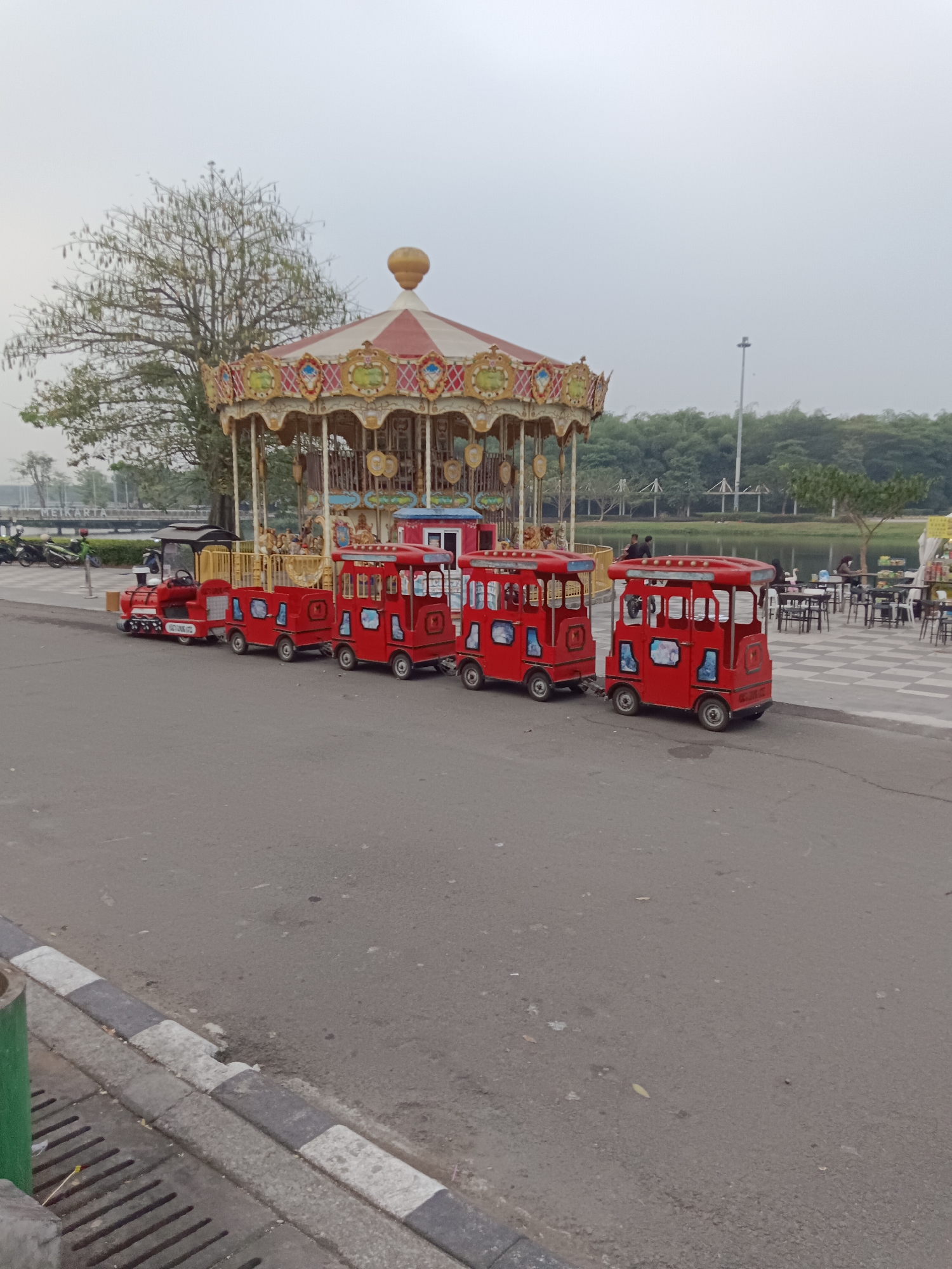 In Jayamukti, Indonesia, a small red train with several passenger cars is parked on an asphalt road. The train is positioned in front of a carousel, which has a decorative roof with red and white stripes and ornate golden trim. Behind the carousel, a body of water is visible, bordered by a line of trees and a clear sky. In the foreground, the road has a gray and white striped curb and a drain. To the left of the train, several motorcycles are parked on a paved walkway. People are visible in the background, seated at tables near the water, and some are walking on the checkered pavement. The overall scene appears to be an outdoor recreational area.
