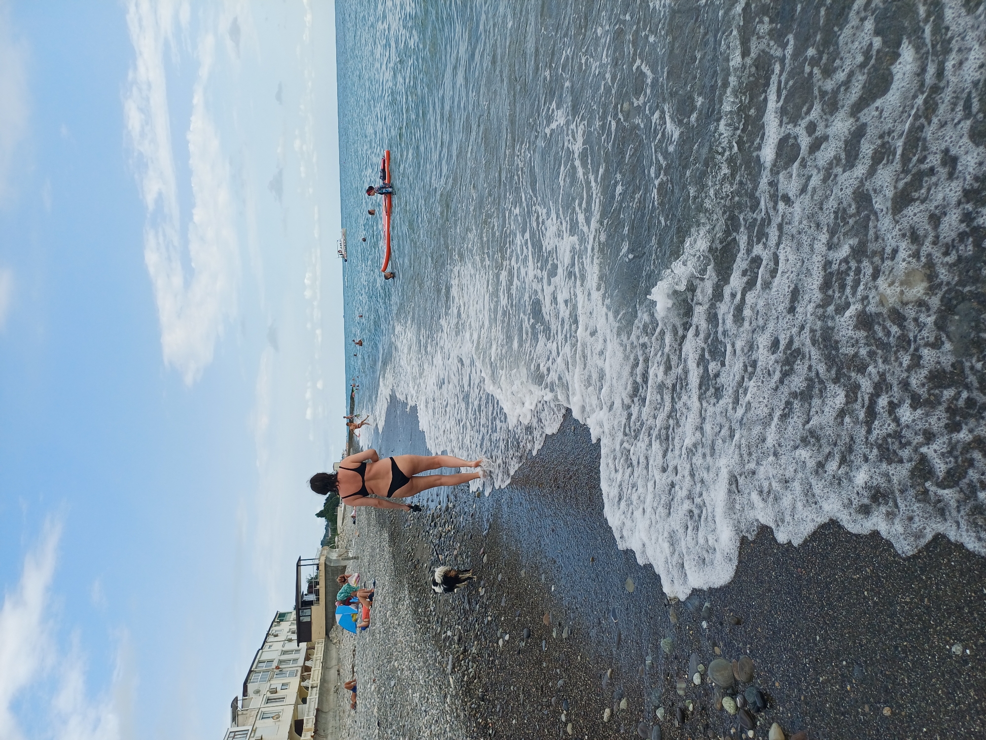 This image depicts a beach scene on a cloudy day, likely in Sochi, Russia, given the architectural style of the buildings in the background which are characteristic of the Black Sea coast. In the foreground, a woman wearing a black bikini walks along the pebble beach, accompanied by a small black and white dog on a leash. Gentle waves lap at the shore, creating white foam. Further out in the sea, several people are visible enjoying the water. One person is on an orange inflatable raft, and another is swimming nearby. In the distance, a boat cruises on the horizon. The beach is lined with buildings that appear to be hotels or apartment complexes, with some people sunbathing or relaxing on the shore. The overall atmosphere is one of a leisurely summer day at the seaside.