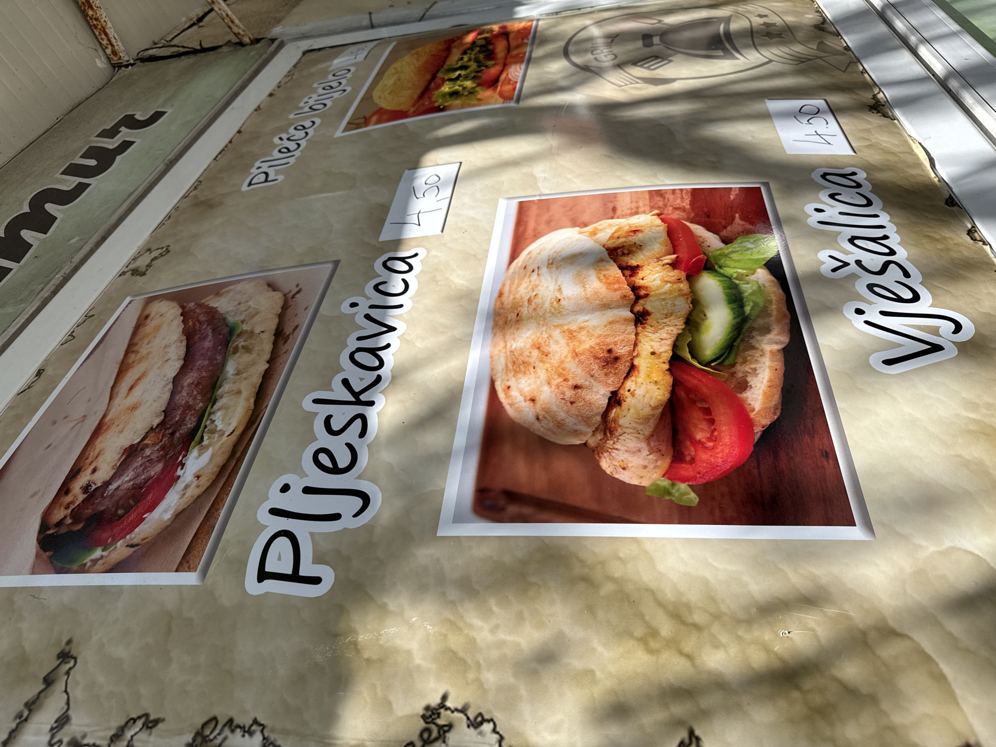 The image shows a close-up of a food menu board, likely attached to a food stall or kiosk. The menu advertises several dishes, including "Pljeskavica" and "Vješalica", both priced at 4.50 (likely Euros). The menu items are depicted with photos of the food, featuring grilled meat in pita bread with vegetables like tomatoes and cucumbers. The background of the menu board has a marble-like texture. The location is likely in Herceg Novi, Montenegro, given the food names and currency. There are no people visible in the image. The scene is well-lit and focuses primarily on the menu offerings.