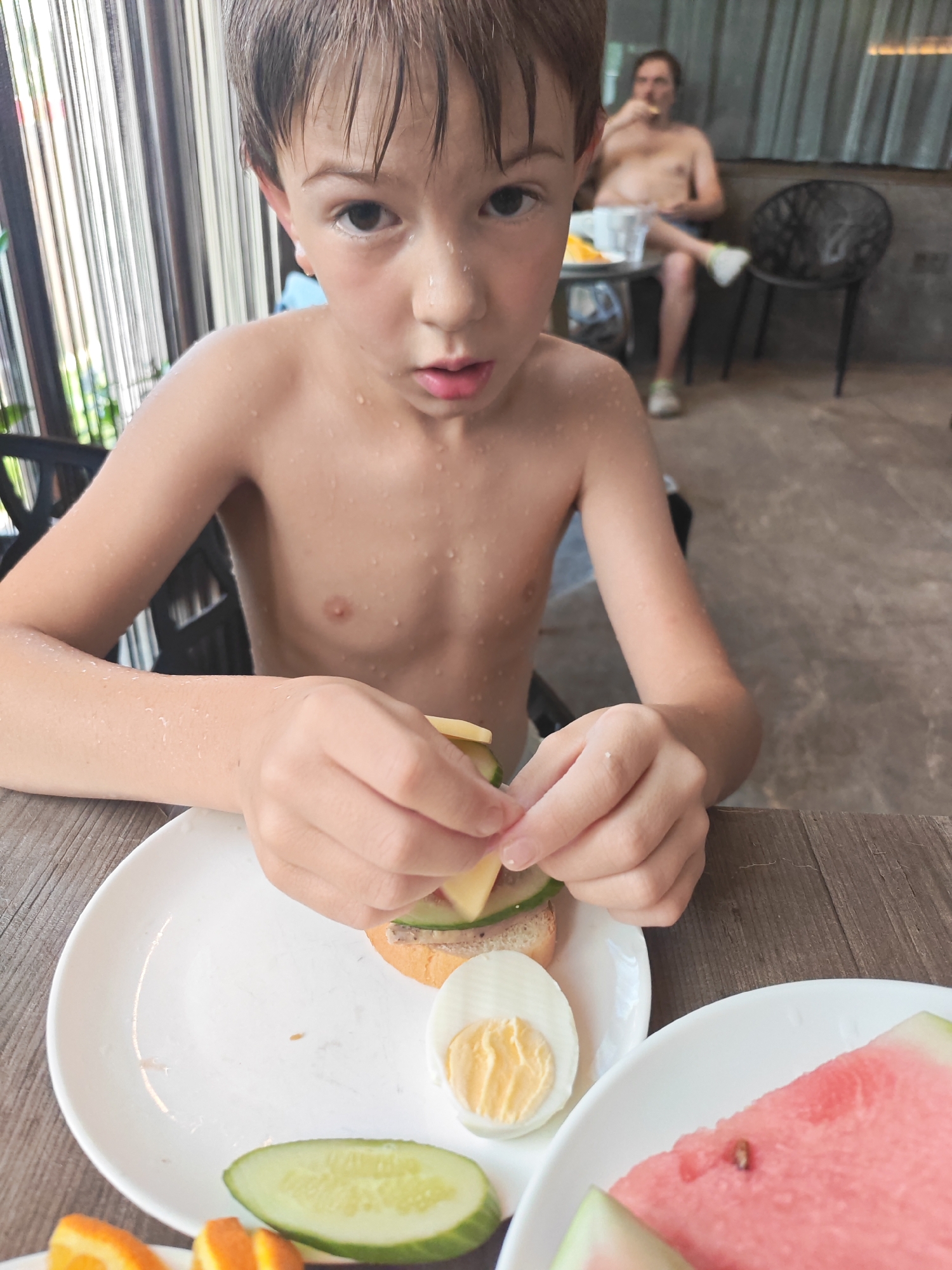 A young boy with wet hair and skin is making a sandwich at a table. He is holding a piece of cheese and placing it on a piece of bread with sliced cucumber and what appears to be a spread. On the plate in front of him are a hard-boiled egg, sliced cucumber, and a slice of watermelon. In the background, an adult male is sitting at another table, eating. The scene appears to be outdoors, possibly at a cafe or restaurant, with a patterned curtain in the background. The overall mood seems relaxed and casual. The time of day is likely daytime, given the lighting. There is no visible text in the image.
