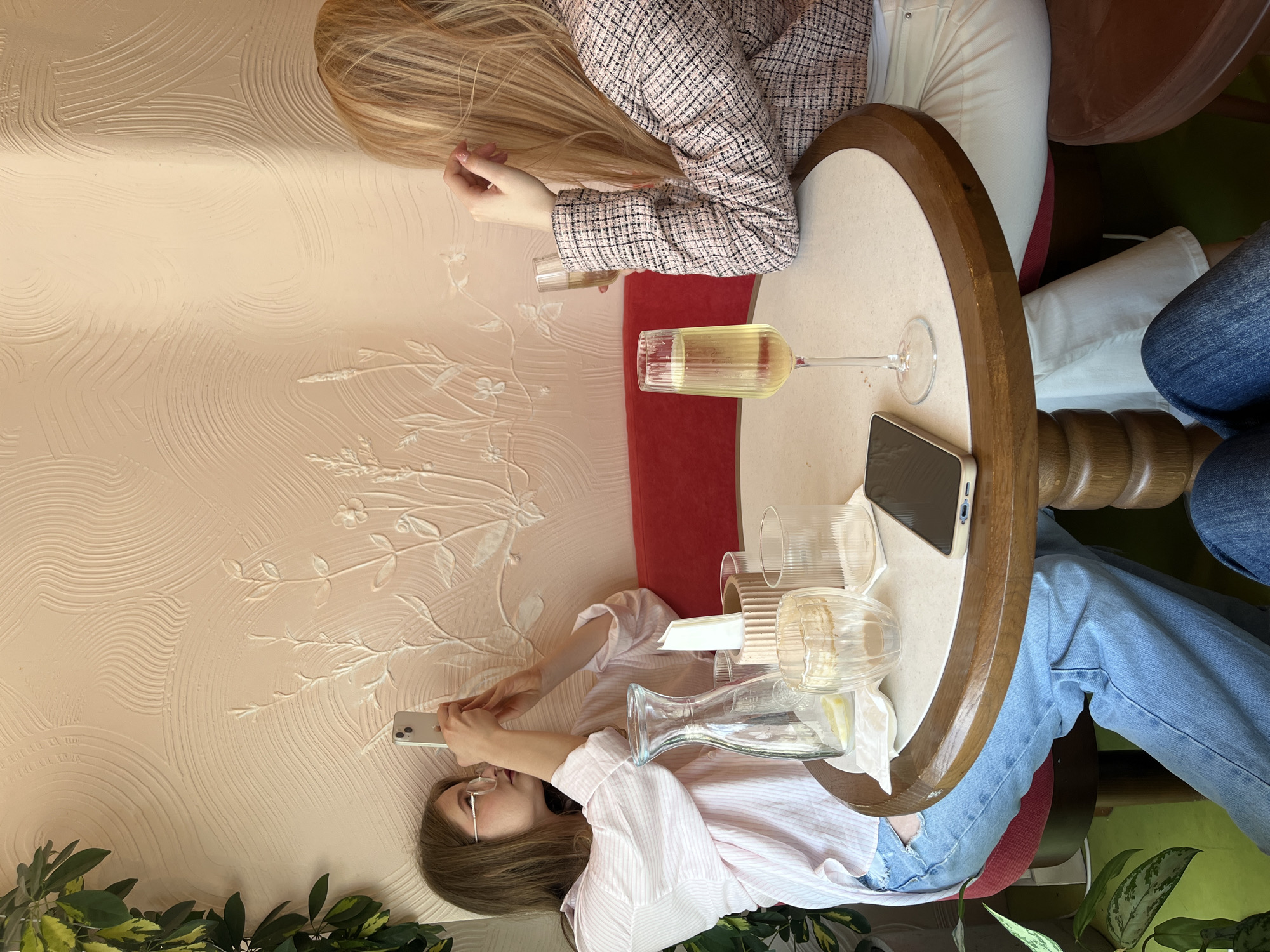 The image shows two young women sitting at a round table in a cafe. The woman on the left is wearing a pink and white striped shirt and blue jeans. She has brown hair and is wearing glasses. She is holding up her phone and taking a picture of the textured pink wall behind her. On the table in front of her is a glass carafe of water with lemon slices, two clear glasses, and a napkin holder. The woman on the right has blonde hair and is wearing a pink and black tweed jacket and white pants. She is holding a champagne flute with a light-colored drink. Another champagne flute is on the table in front of her. The wall behind them has a decorative, textured pattern with raised floral motifs. A green plant with variegated leaves is visible on the left side of the frame, and a red banquette runs along the wall. The floor is a lime green color. The overall atmosphere appears casual and social.