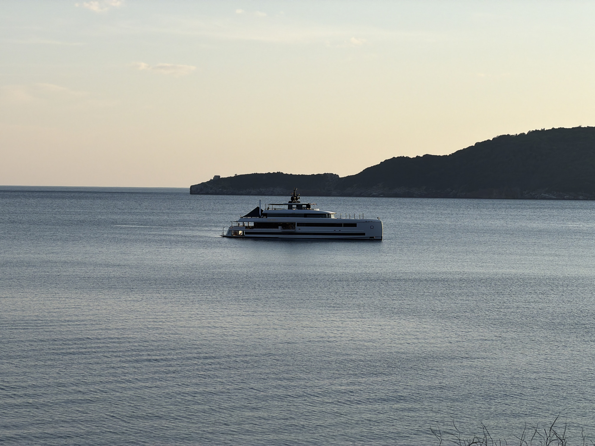 A daytime shot shows a white yacht on calm blue water. In the background, a land mass is visible with a small structure on the left side and a larger landmass on the right. The sky is pale and even. The location is unknown.