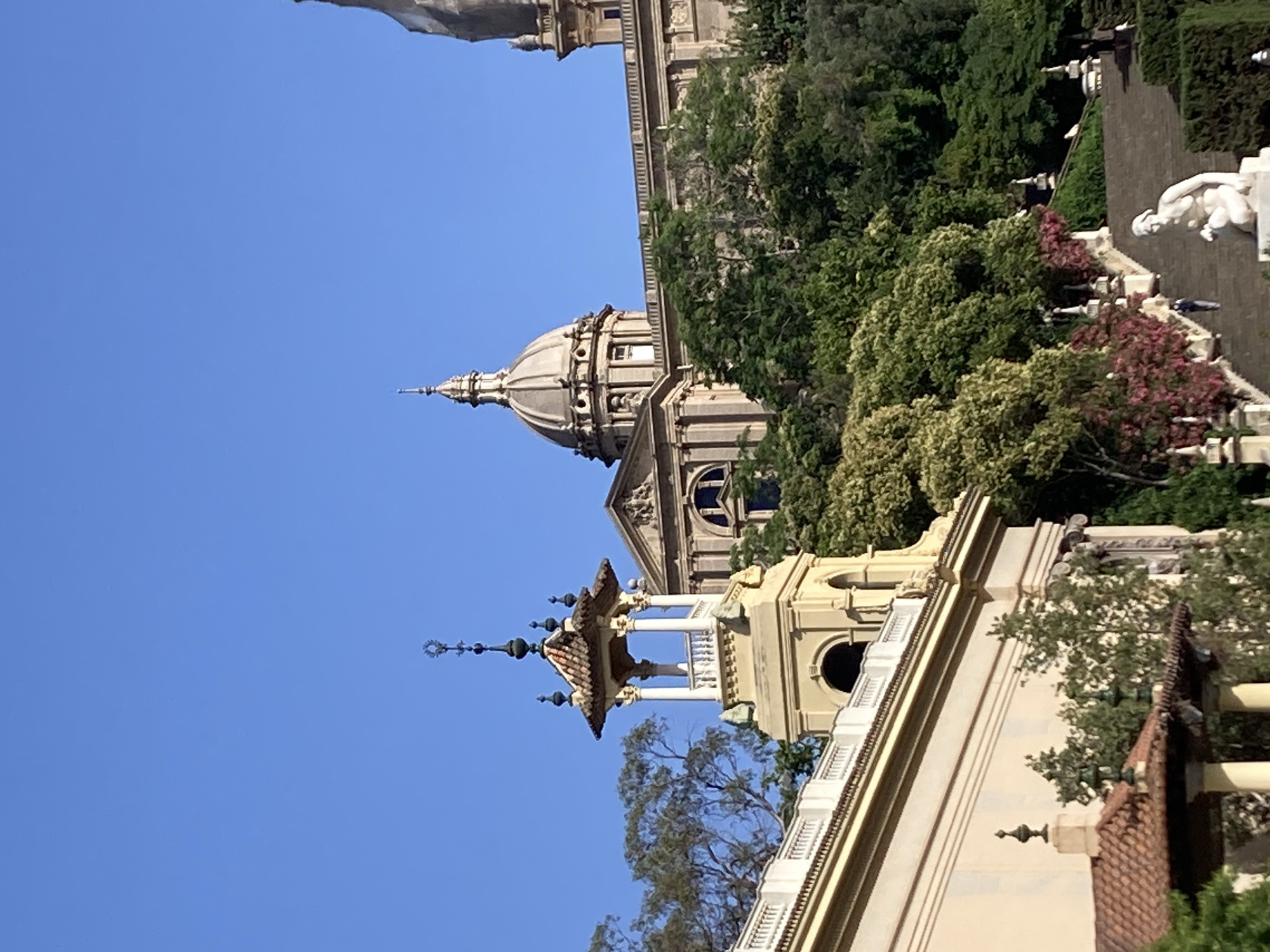 The image shows a view of the Palau Nacional, located on Montjuïc hill in Barcelona, Spain, against a clear blue sky. The Palau Nacional is a large, imposing building with a prominent dome and classical architectural elements, including columns and ornate detailing. In the foreground, a lighter-colored building with a red-tiled roof and a decorative spire is visible. Lush green trees and some flowering bushes are scattered throughout the scene, adding a touch of nature to the architectural landscape. A wide set of stairs leads up to the Palau Nacional, and a few people can be seen walking on them. A white sculpture of a reclining nude figure is positioned at the bottom of the stairs. The overall impression is one of grandeur and historical significance.