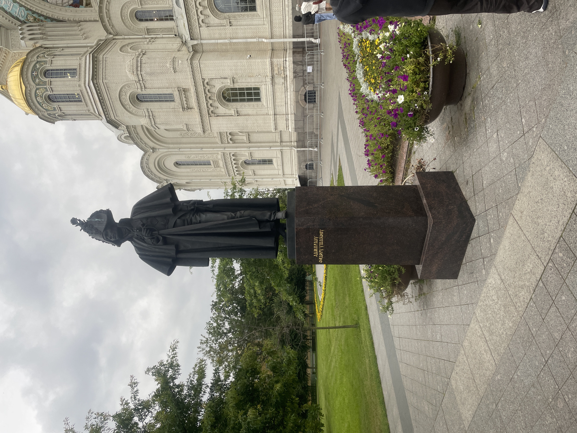 The image depicts a statue of a man in historical military attire, standing on a granite pedestal. The inscription on the pedestal reads "АДМИРАЛУ ФЕДОРУ УШАКОВУ" (To Admiral Fyodor Ushakov). Behind the statue, a large, ornate white building with a golden dome is visible, identified as the Naval Cathedral of Saint Nicholas in Kronstadt, Russia. The foreground features a paved walkway and planters filled with purple and yellow flowers. The sky is overcast. Two individuals are partially visible on the right side of the frame, near the building.