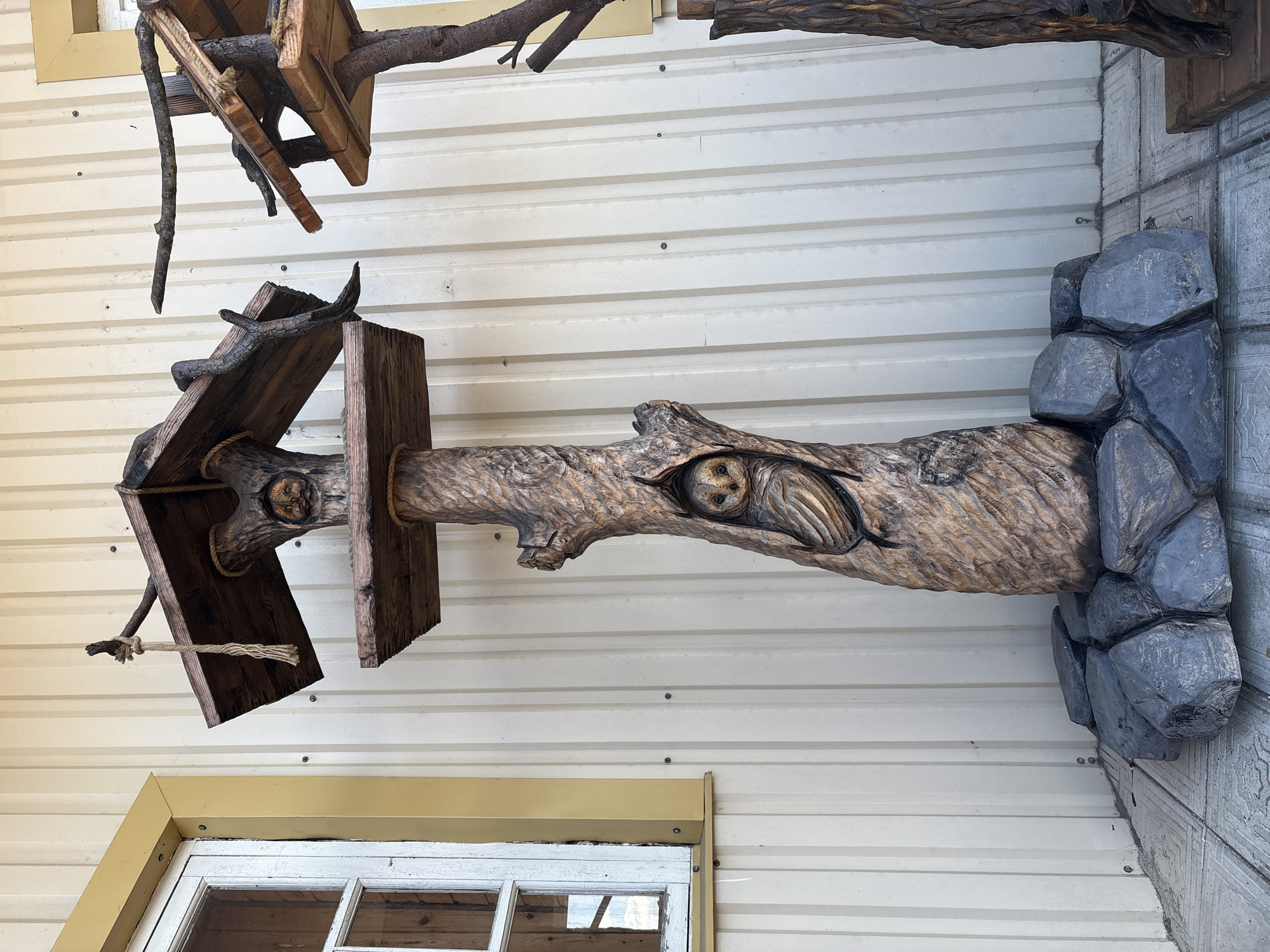 The image shows a carved wooden sculpture of a tree with two owls in carved niches, placed outdoors against a building with corrugated siding. The sculpture is positioned on a tiled surface and surrounded by a cluster of grey, rock-like objects at its base. A window with a yellow frame is visible to the left of the sculpture. The overall setting appears to be a commercial or public space.