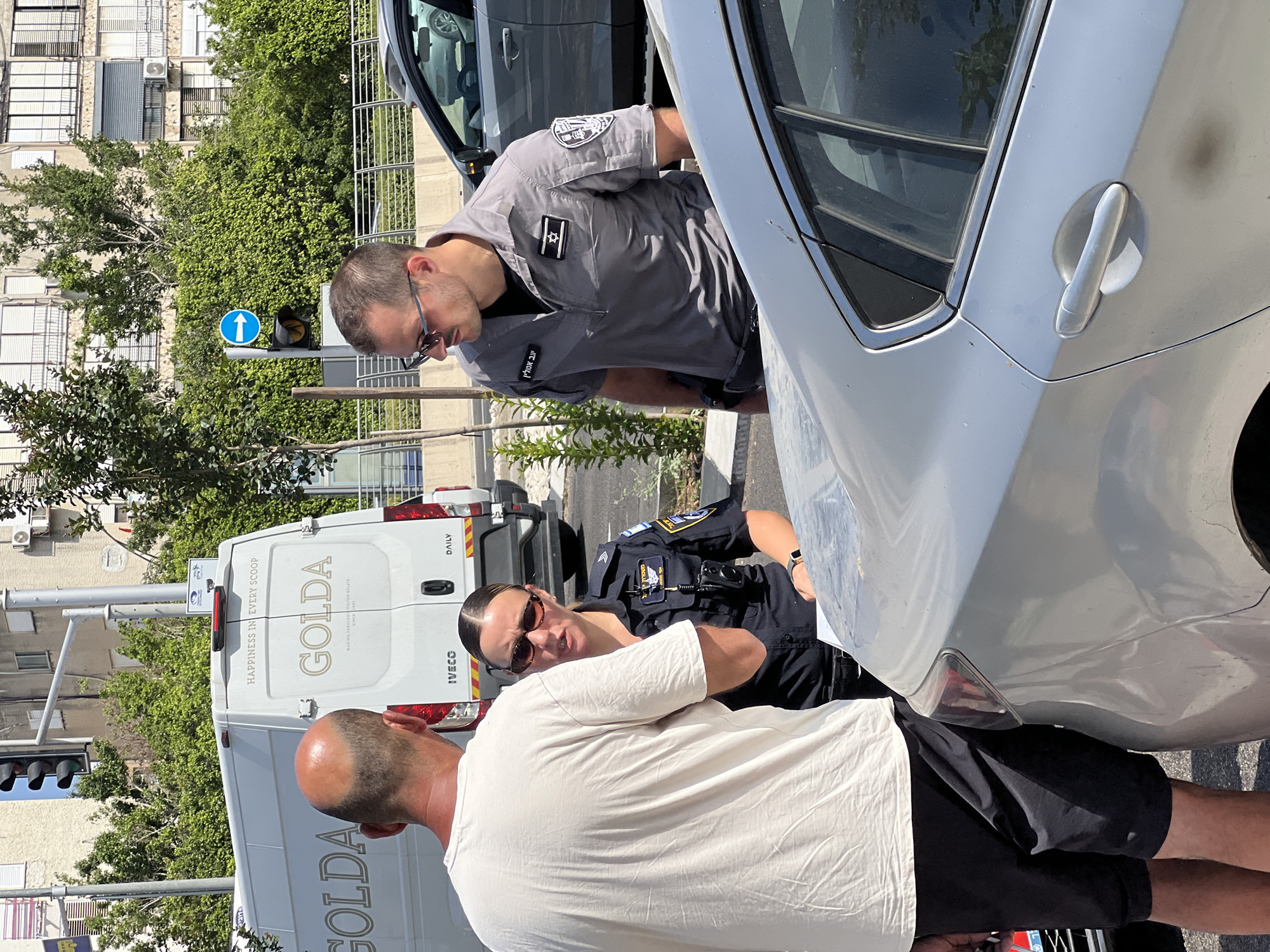 The image depicts an outdoor scene in Kiryat Bialik, Israel, likely on a street. In the foreground, a silver car is partially visible. Three individuals are engaged in a conversation next to the car. One individual, a man wearing a light-colored t-shirt and dark shorts, is facing away from the camera. To his right, a female police officer in uniform is speaking with him, wearing sunglasses and a dark uniform with patches. On the other side of the officer, a male police officer, also in uniform, is standing and looking towards the conversation. Behind the individuals, a white van with "GOLDA" branding is parked. The van also displays the text "HAPPINESS IN EVERY SCOOP" and "MAKING FABULOUS ITALIAN GELATO SINCE 2009." In the background, buildings and lush green trees are visible, along with a traffic light and a blue directional sign with a white arrow. The overall lighting suggests a bright, sunny day.