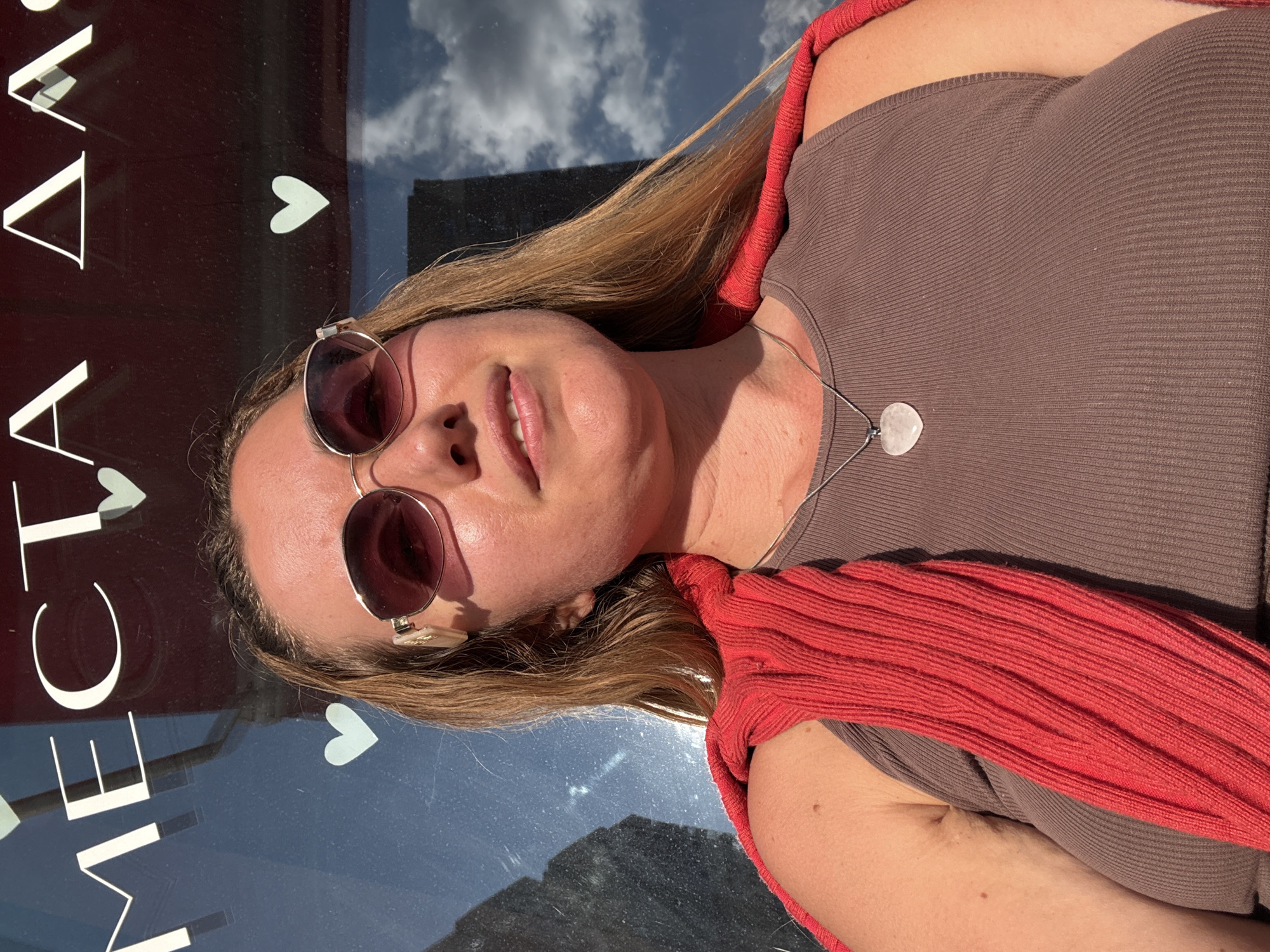 A medium shot shows a woman with long blonde hair and sunglasses wearing a brown tank top and a red sweater draped over her shoulders. She is looking slightly up and to her right, with a faint smile. A silver necklace with a heart-shaped pendant rests on her chest. In the background, a dark, reflective surface displays white lettering and light blue hearts. The lettering appears to be in a foreign language, with some letters visible as "MECTA AA." The reflective surface also shows a reflection of the sky with clouds and a dark building. The lighting suggests it is daytime, possibly outdoors.